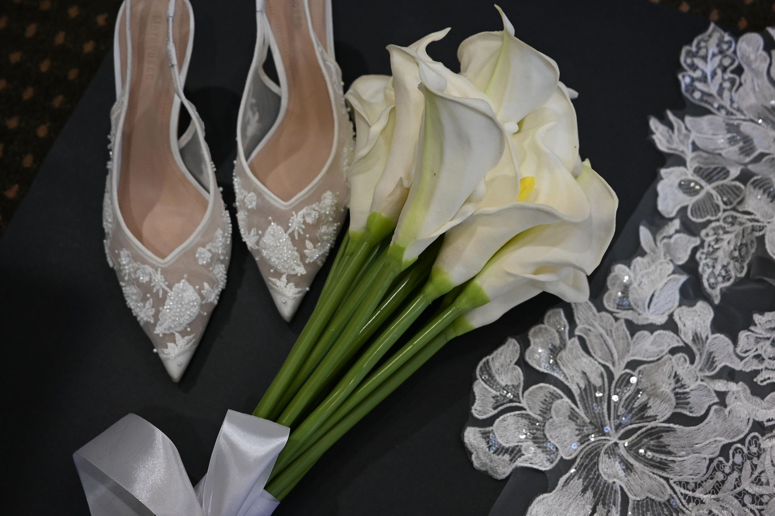 Elegant white calla lilies and lace arranged with bridal shoes against a dark background for a wedding detail shot.