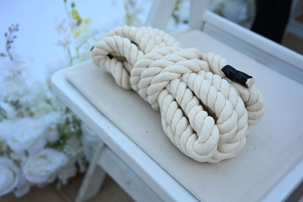 A coiled white cotton rope with intricate knots sits on a white table near floral decorations, used in Christian knot ceremonies at wedding