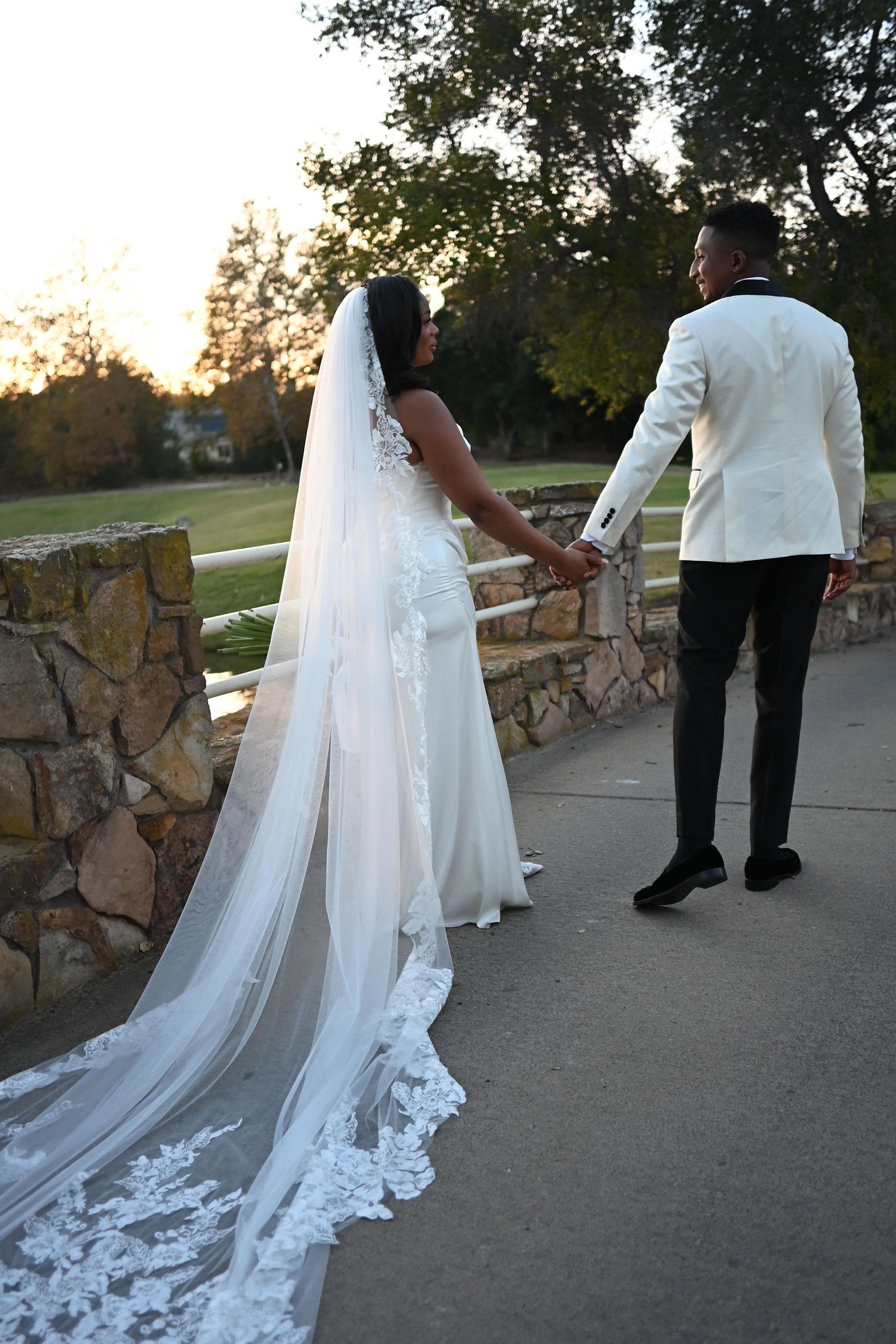 A bride in a flowing white dress with long lace veil stands with her groom at sunset on a stone wall pathway.