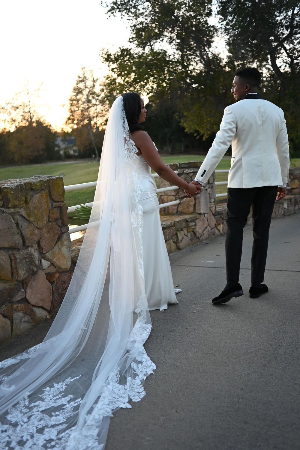 A bride in a flowing white dress with long lace veil stands with her groom at sunset on a stone wall pathway.