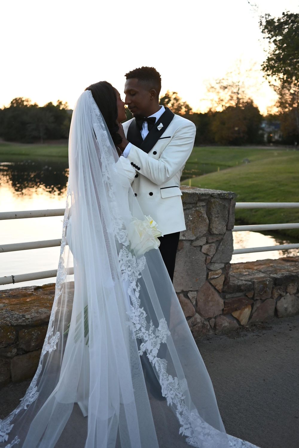 A romantic sunset wedding photoshoot with a couple embracing by a stone wall and white fence in a scenic outdoor setting.