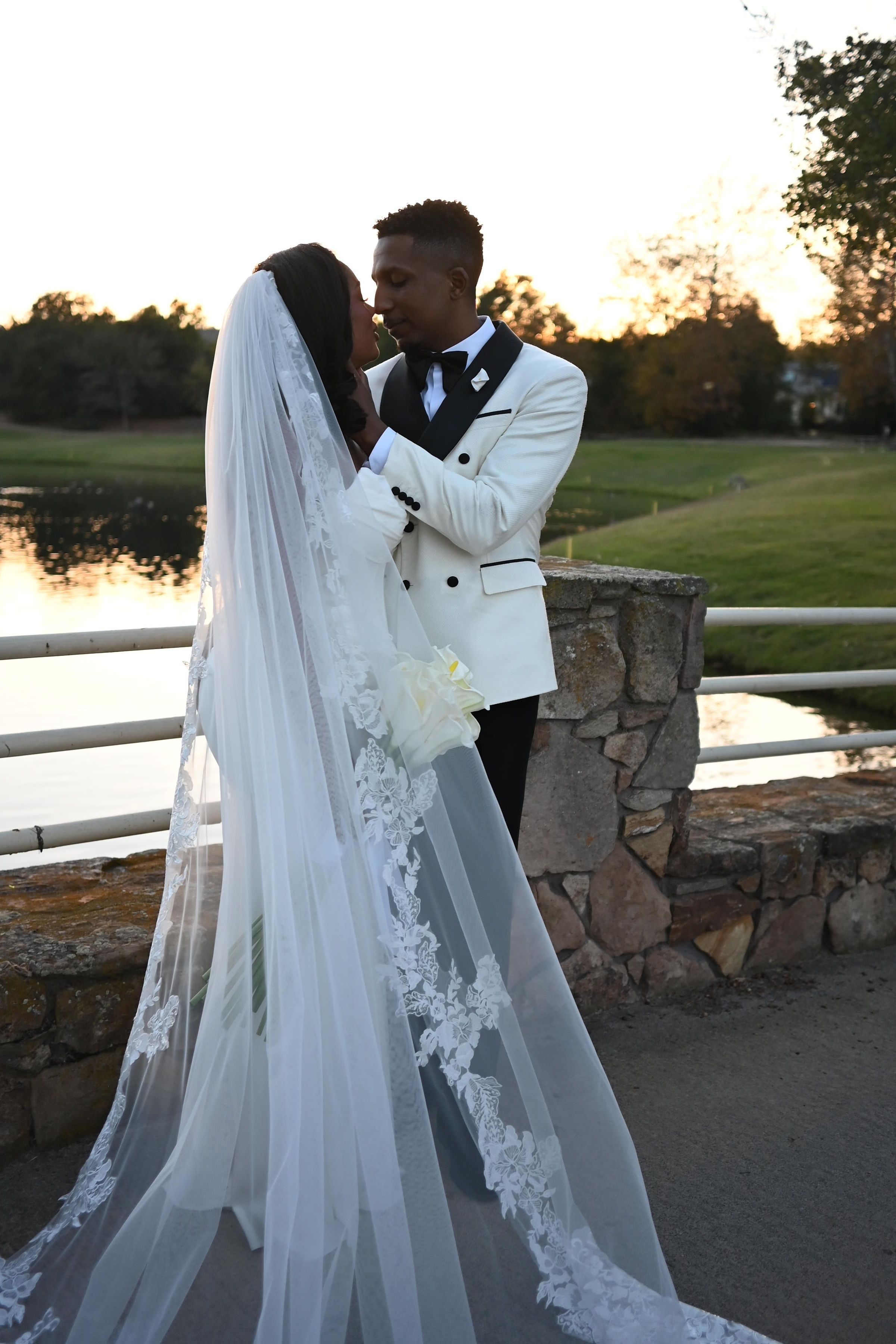 A romantic sunset wedding photoshoot with a couple embracing by a stone wall and white fence in a scenic outdoor setting.