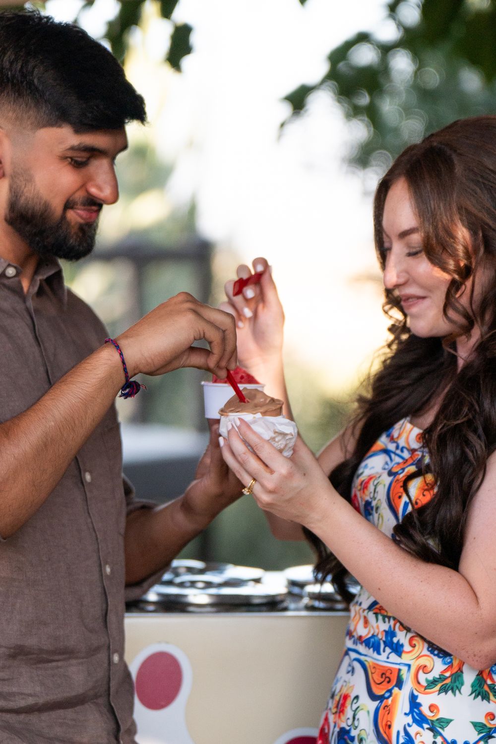 A couple shares a playful moment while feeding each other ice cream after their engagement outdoors on a sunny day.