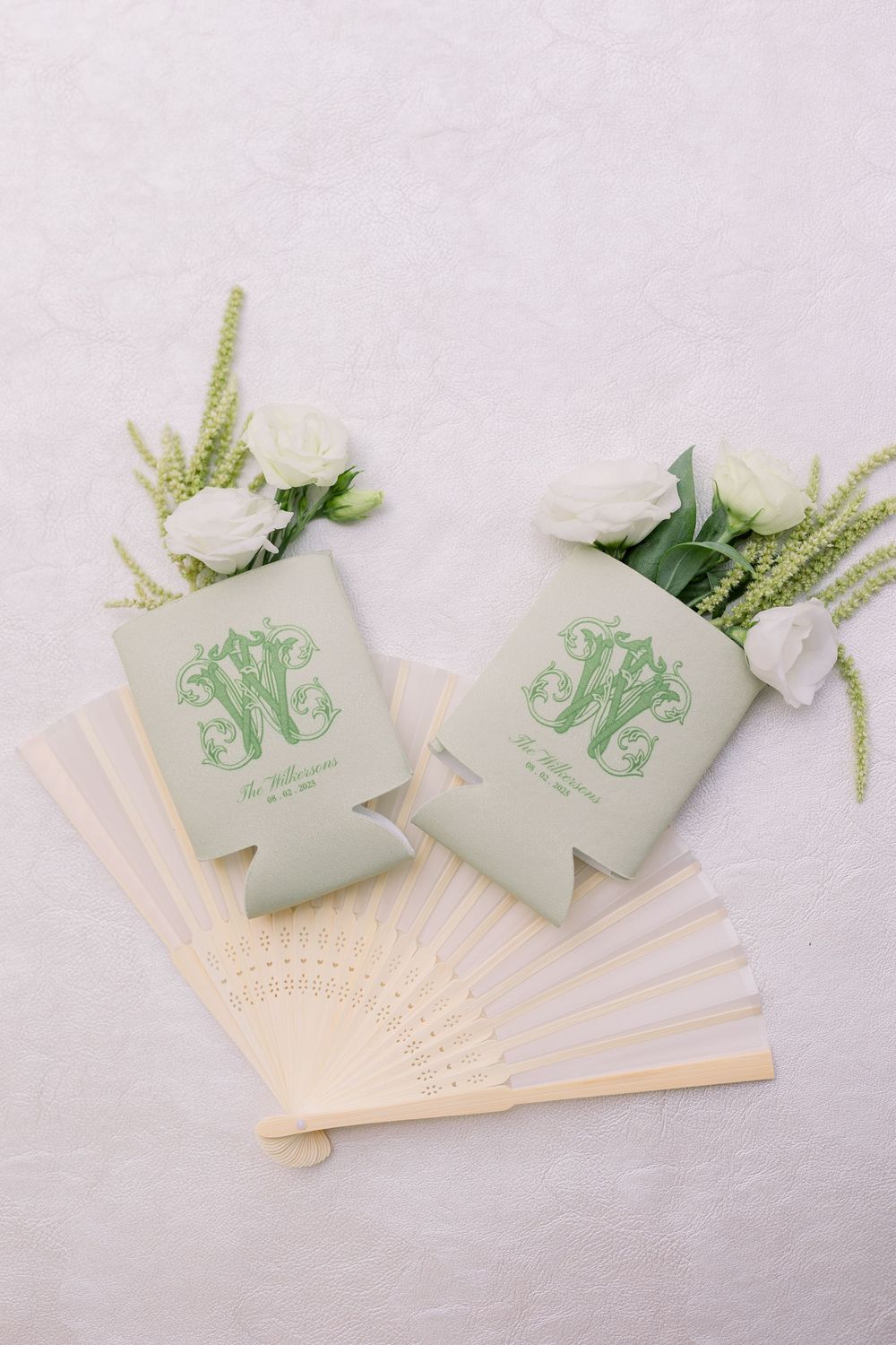 Mint green monogrammed wedding koozies displayed with decorative fans and greenery.