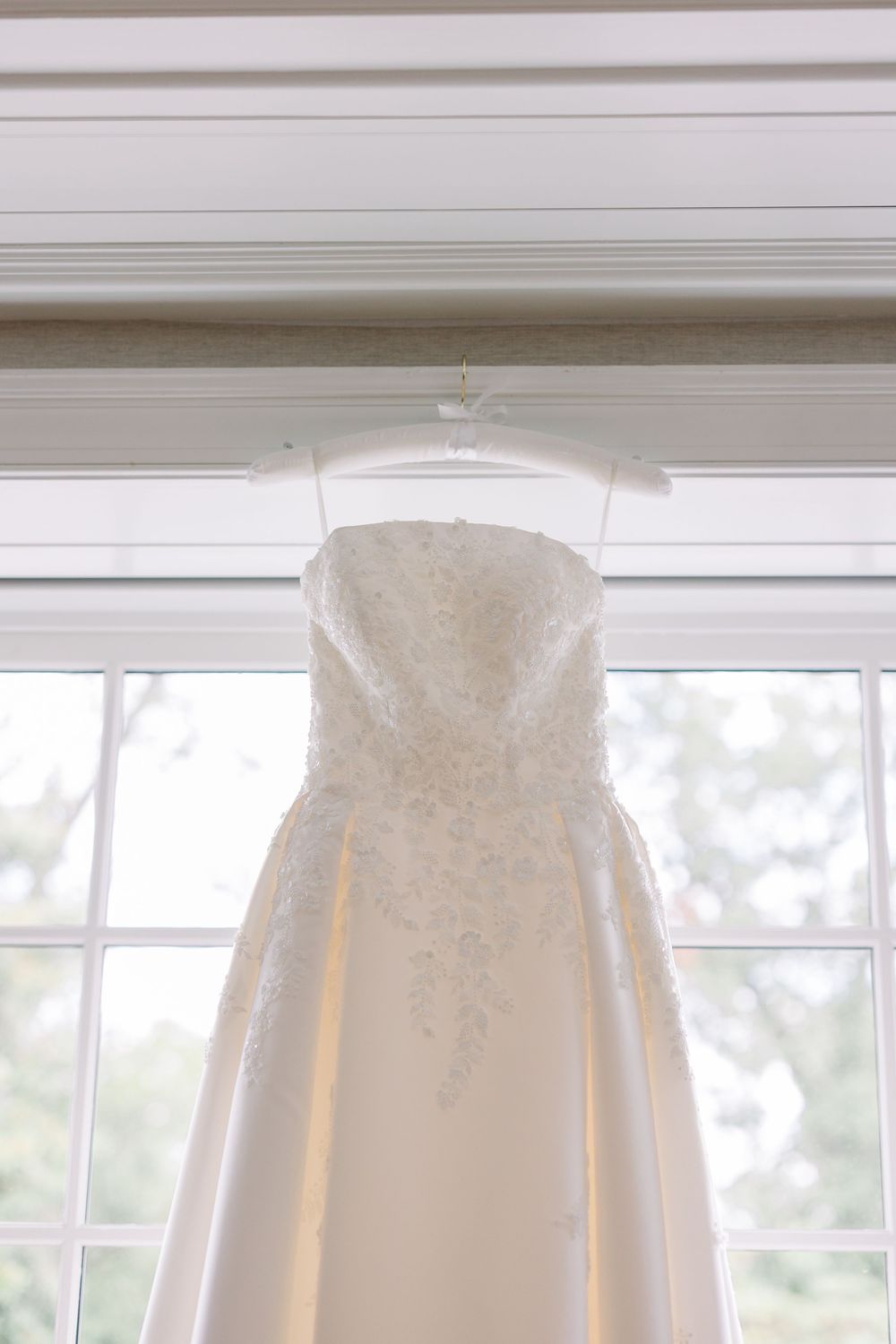 A white wedding dress with lace details hangs elegantly against a window with natural light streaming in.