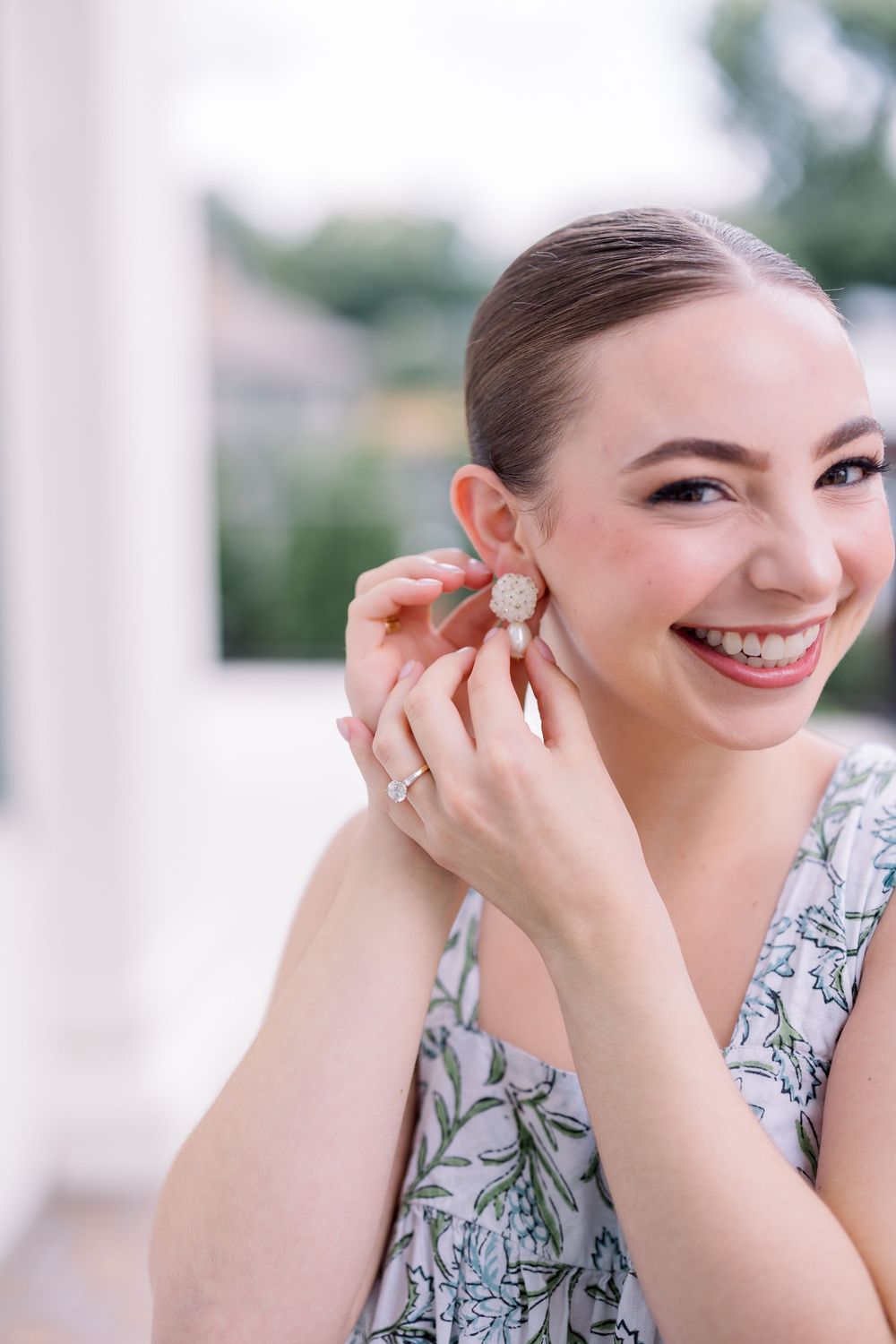 Elegant woman in floral dress smiling while putting on earrings during special occasion.