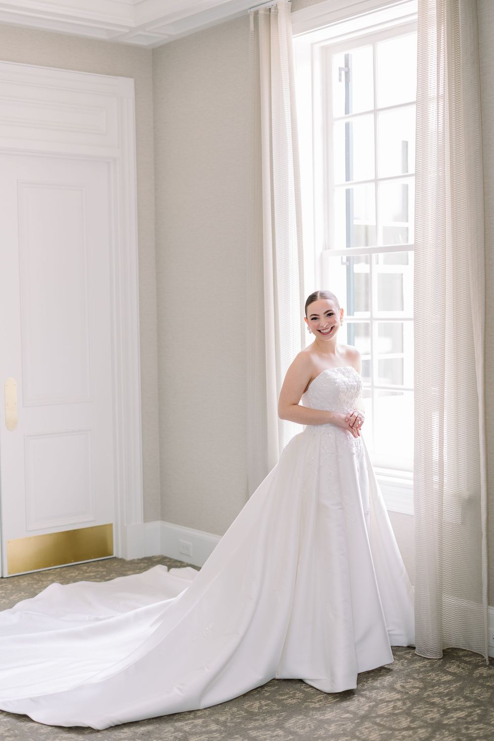 An elegant white wedding gown with a long train photographed in a bright room with large windows.