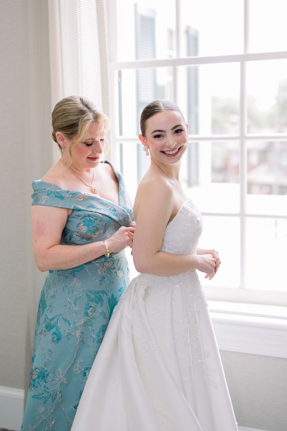 A bride getting dressed in a white gown while someone in a turquoise dress helps with the preparations.