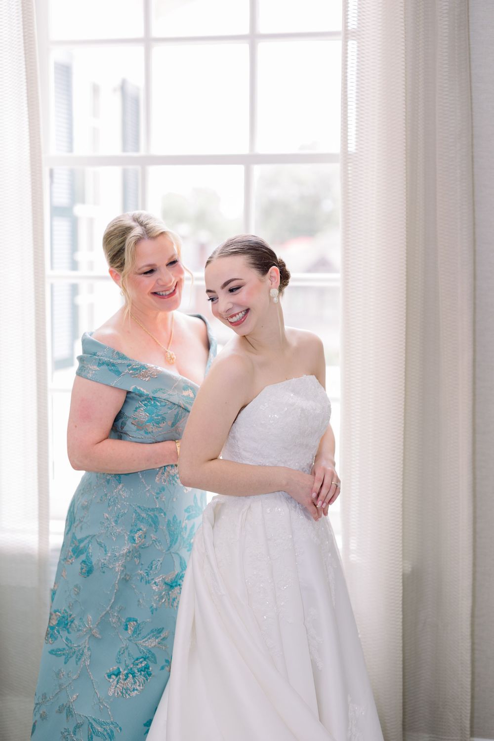 Two people share an emotional moment in a bright room, one wearing a light blue dress and the other in white bridal gown.