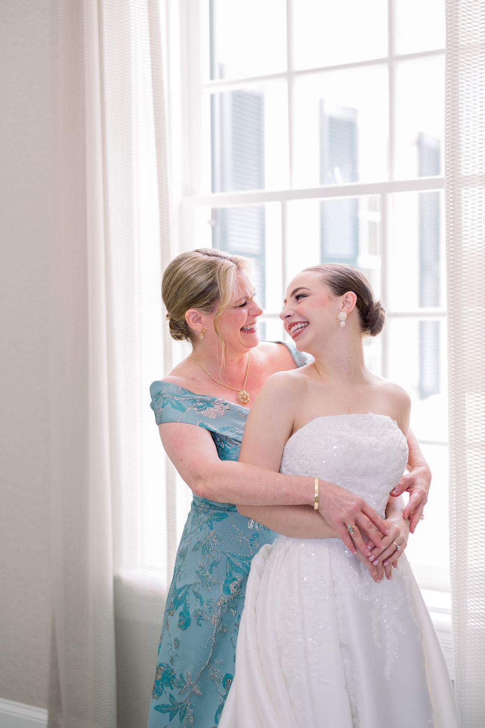 Two women sharing a joyful embrace in a sunlit room with white curtains.