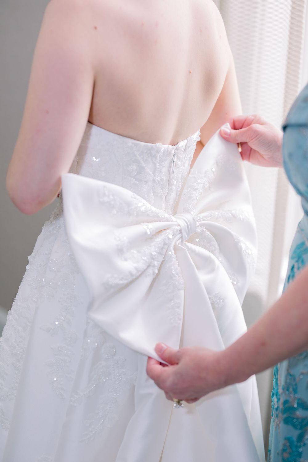 Close-up detail of a white wedding dress bow being tied at the back.
