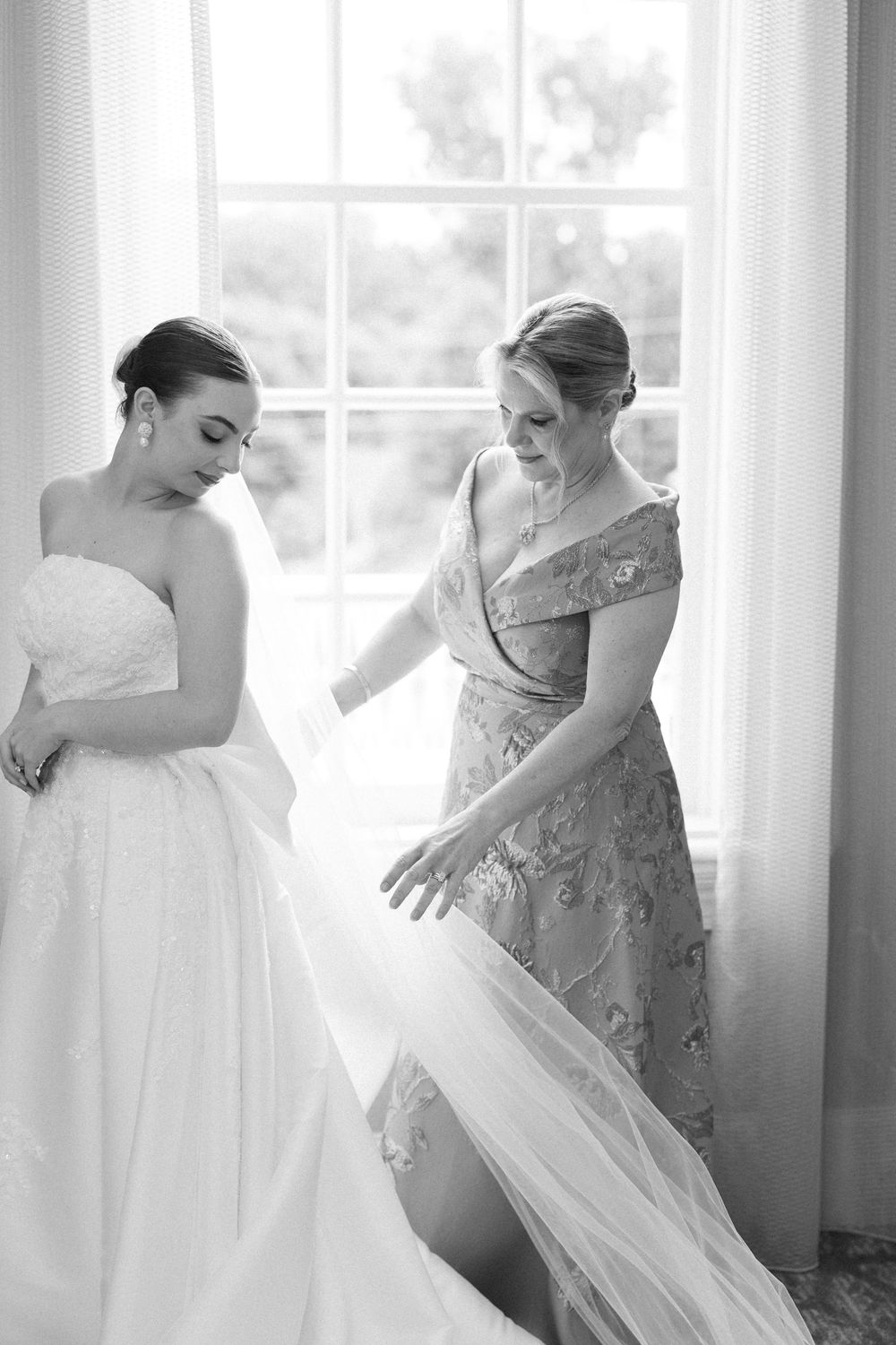Intimate getting ready moment between bride and bridesmaid in flowing dresses by window.