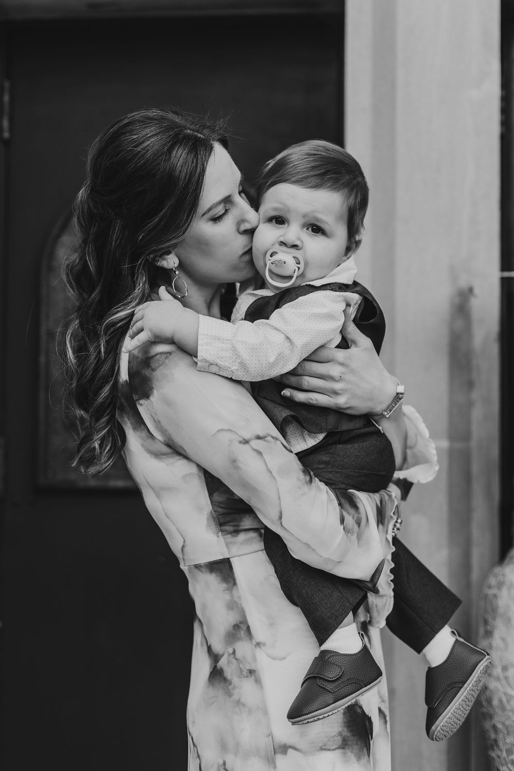 A series of tender black and white moments showing a mother kissing and embracing a baby in formal attire.