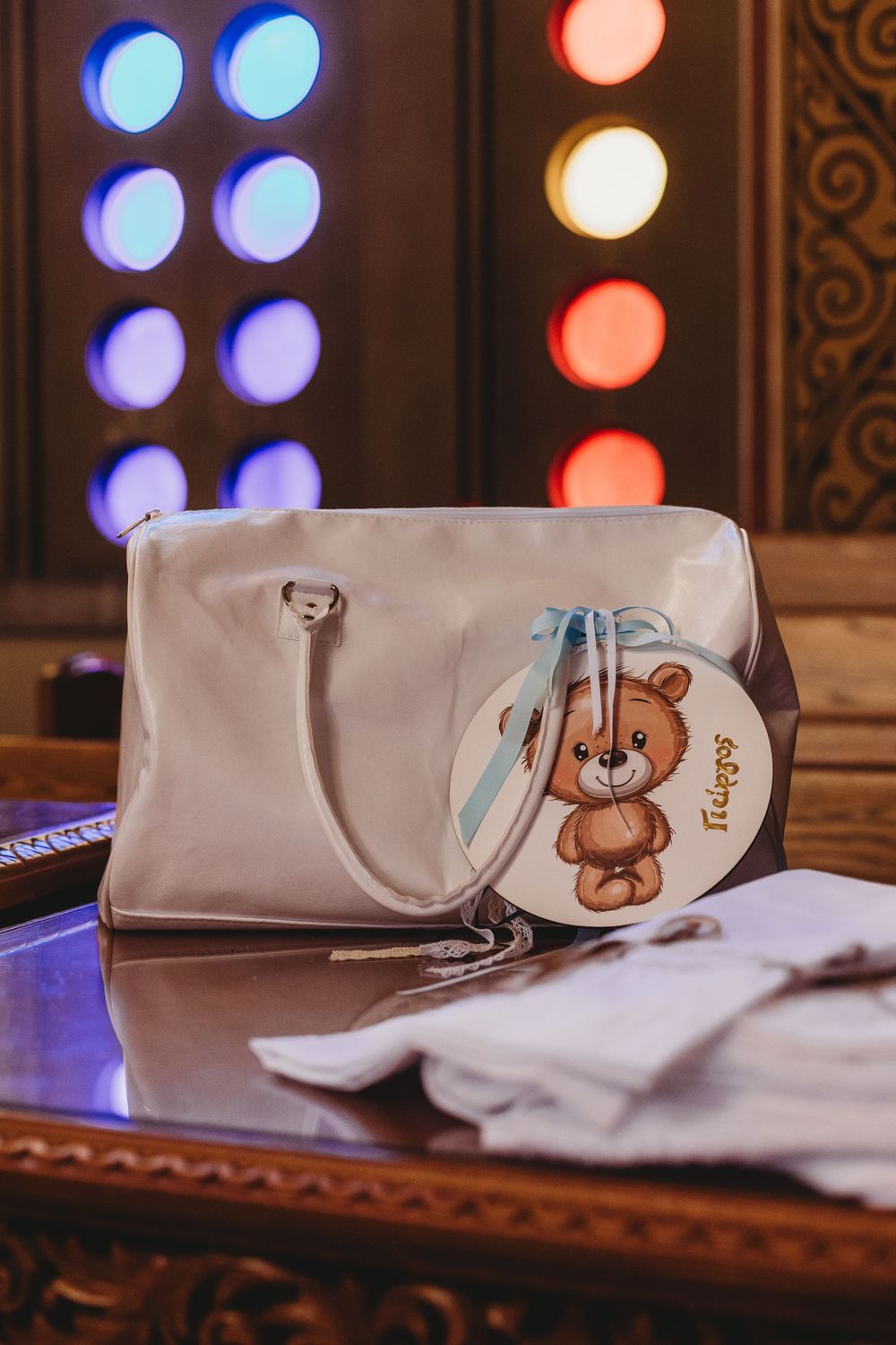 A beige handbag with a cute teddy bear charm sits on a table with blue and red lights in the background.