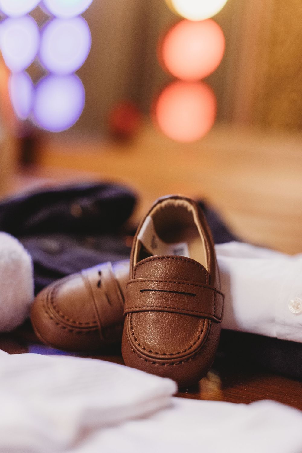 A small brown leather baby loafer shoe photographed against a soft background with bokeh lights.