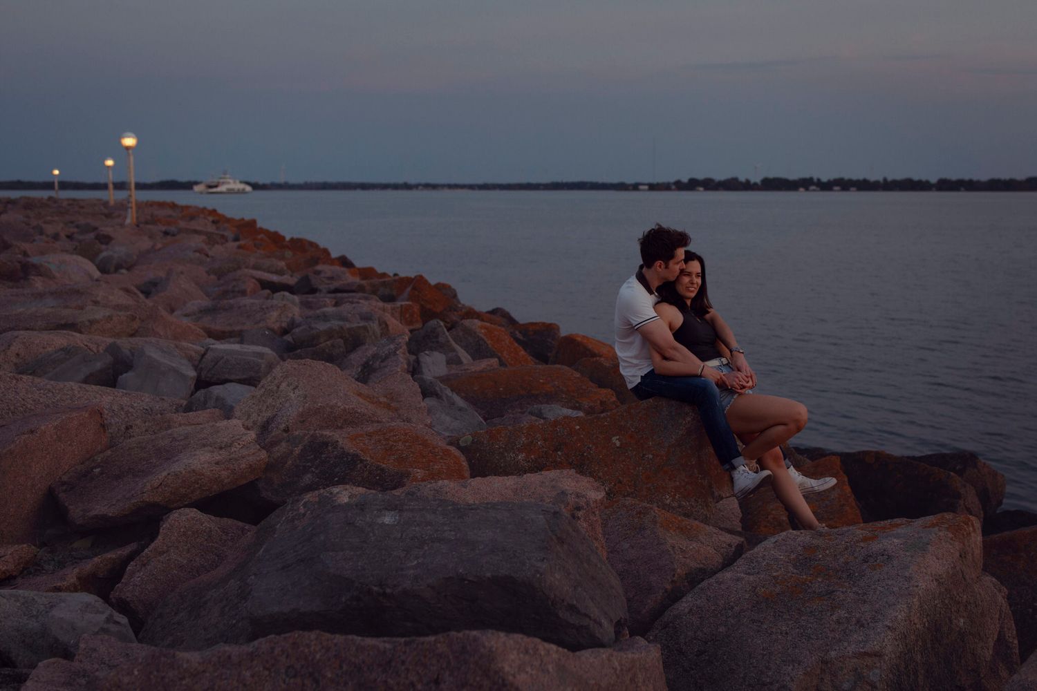 couple sit on the armor rock as the sun sets, the ferry crosses lake ontario behind them.