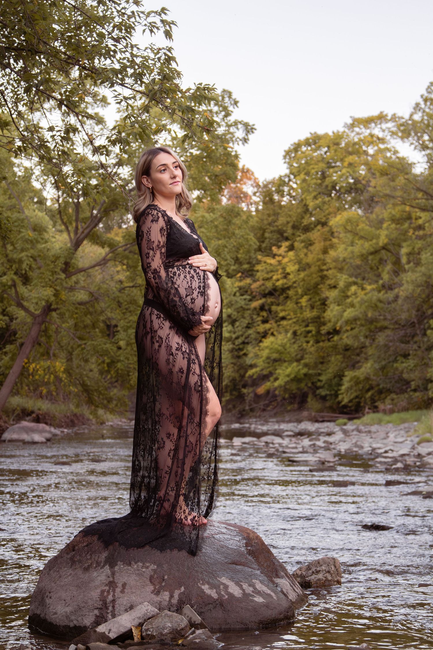 outdoor maternity session mom on rock in the water holding belly in open front lace dress