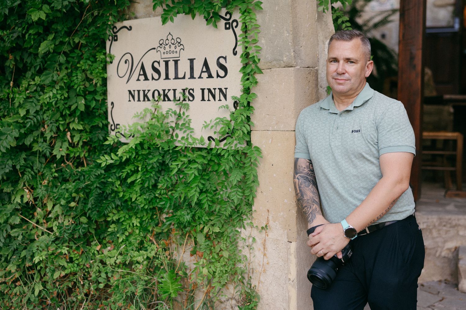 A rustic wooden sign for Vasilias Inn hangs on a stone wall surrounded by lush green foliage.