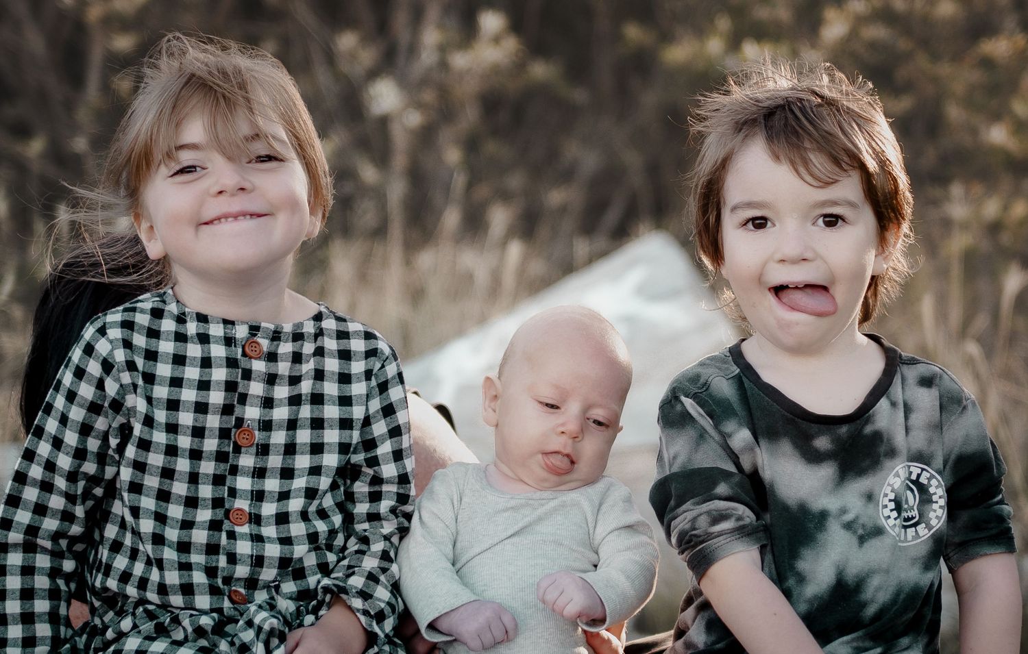 Two young siblings smile outdoors while holding a newborn baby between them in a natural setting.