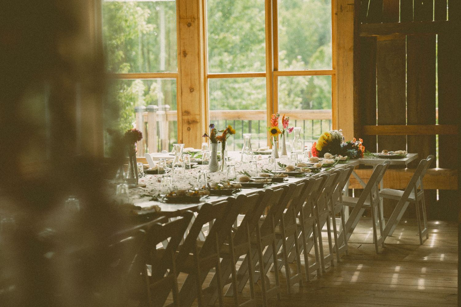 Long rustic wooden tables set for an elegant wedding reception in a barn with natural lighting streaming through windows.