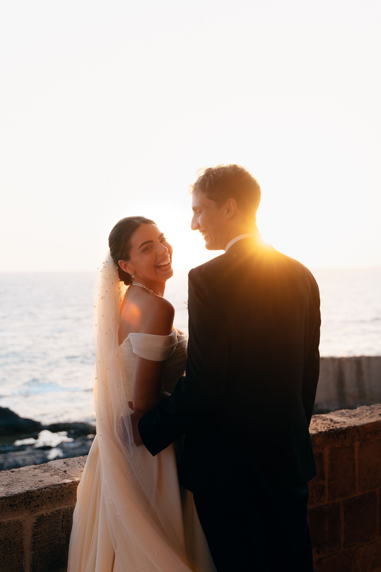 A bride and groom share an intimate moment at sunset by the ocean, silhouetted against the golden light.