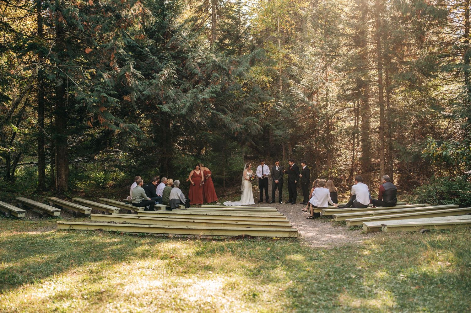 Outdoor wedding ceremony setup in a sunlit forest with wooden bench seating and guests gathered on rustic amphitheater steps.