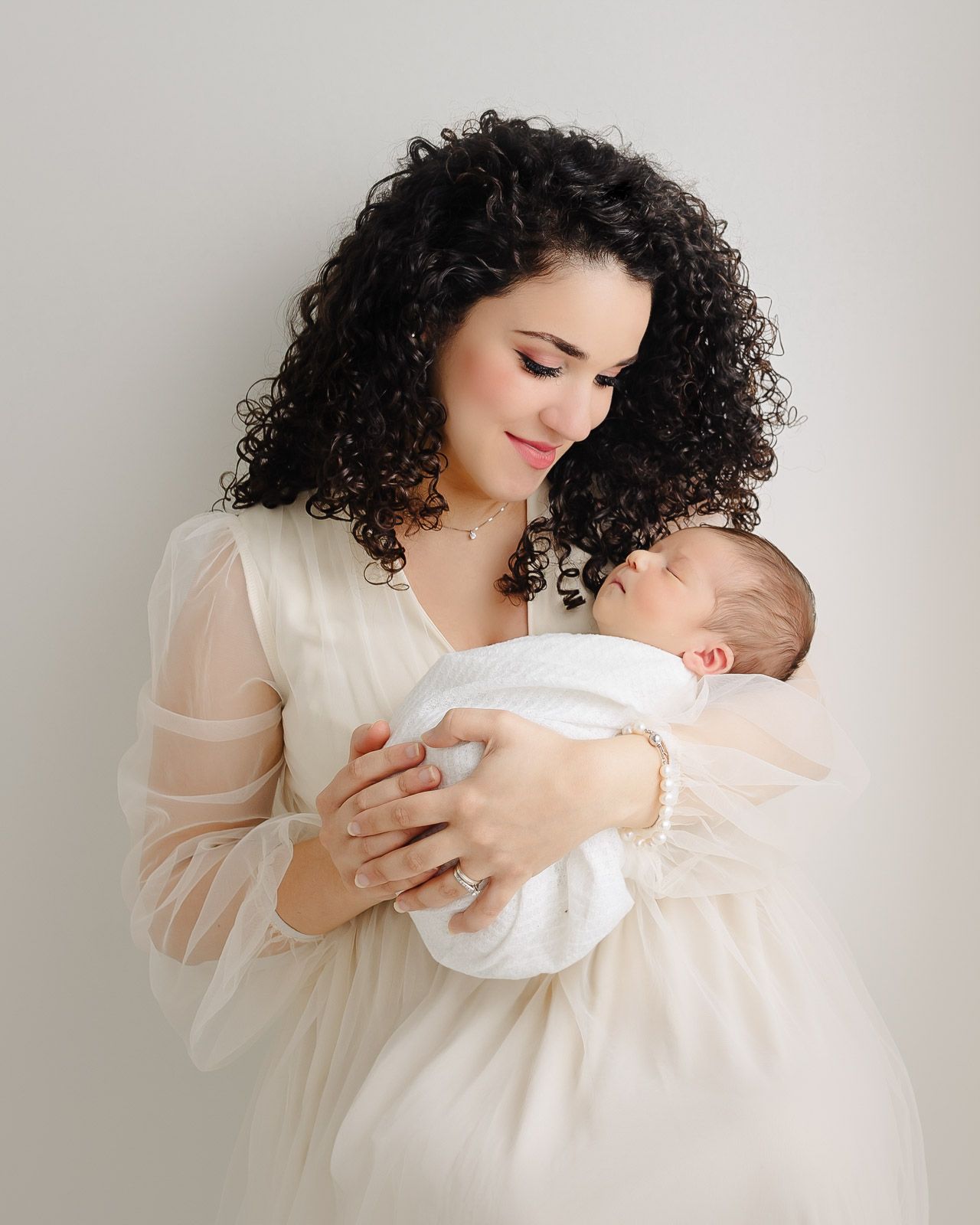 A mother in a cream-colored dress cradles and gazes lovingly at her newborn baby wrapped in white.