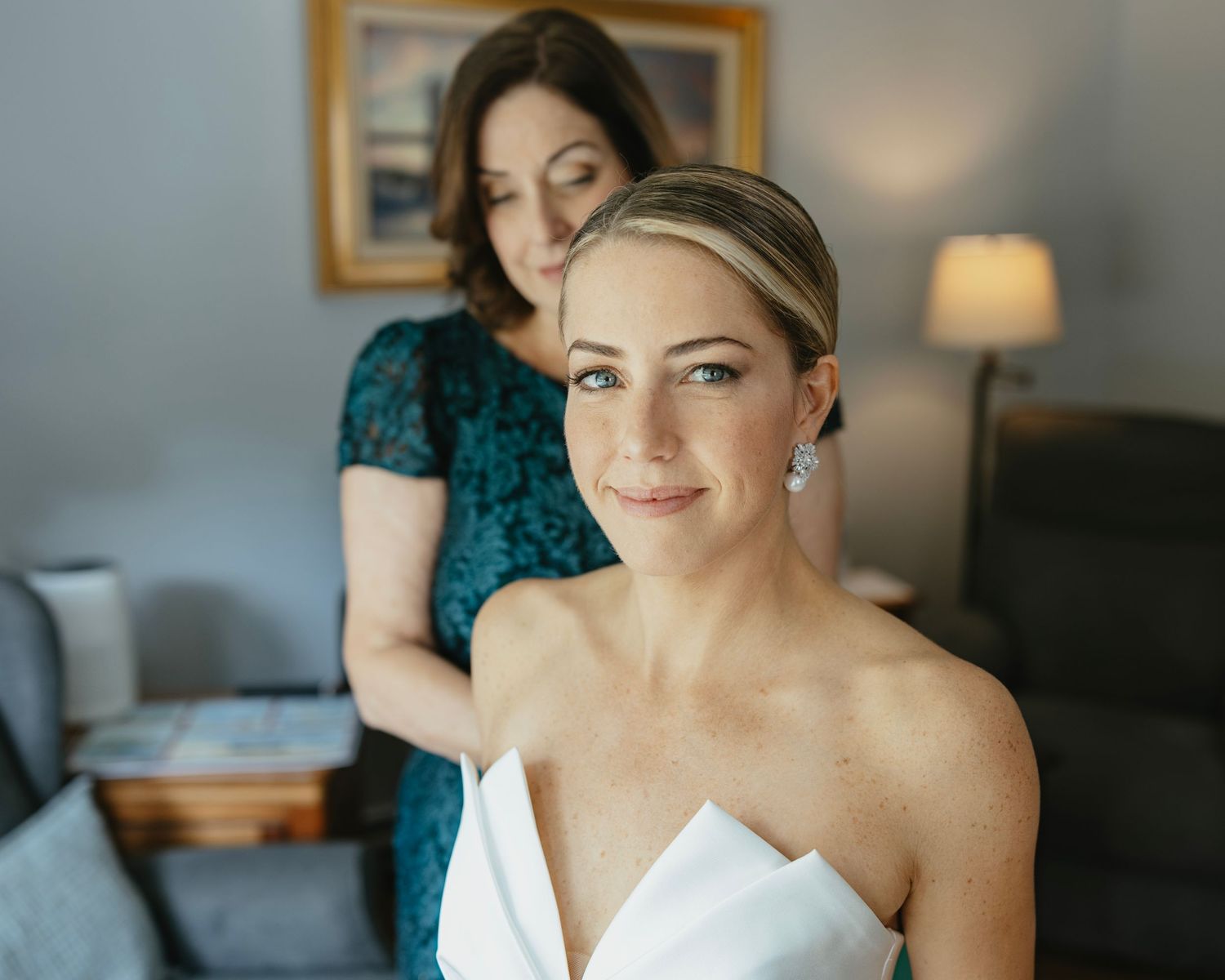 A bride in a white strapless dress gets ready with hair styling assistance in a softly lit room.
