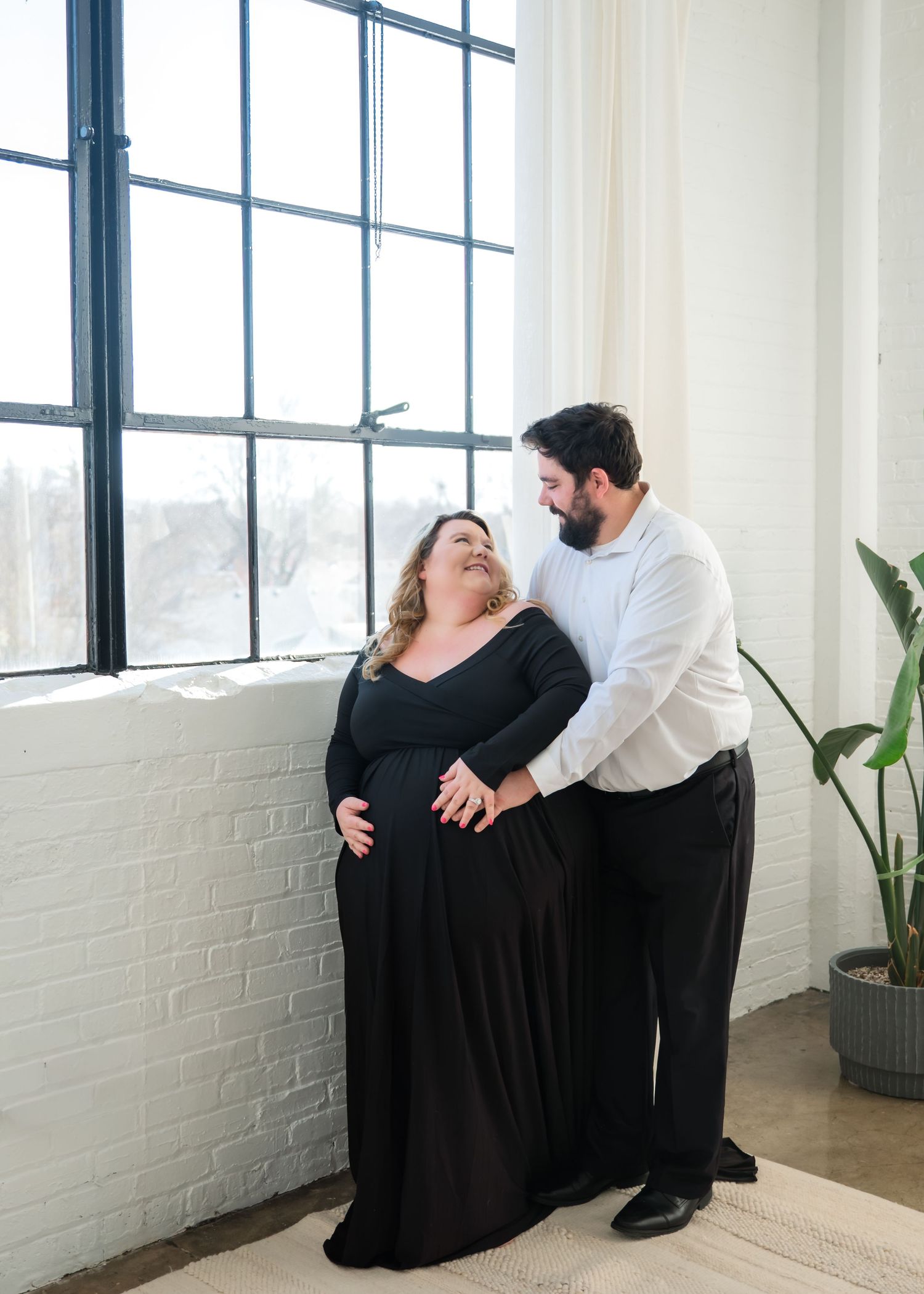 A couple shares a joyful moment together while standing by a large industrial window in a bright white room.