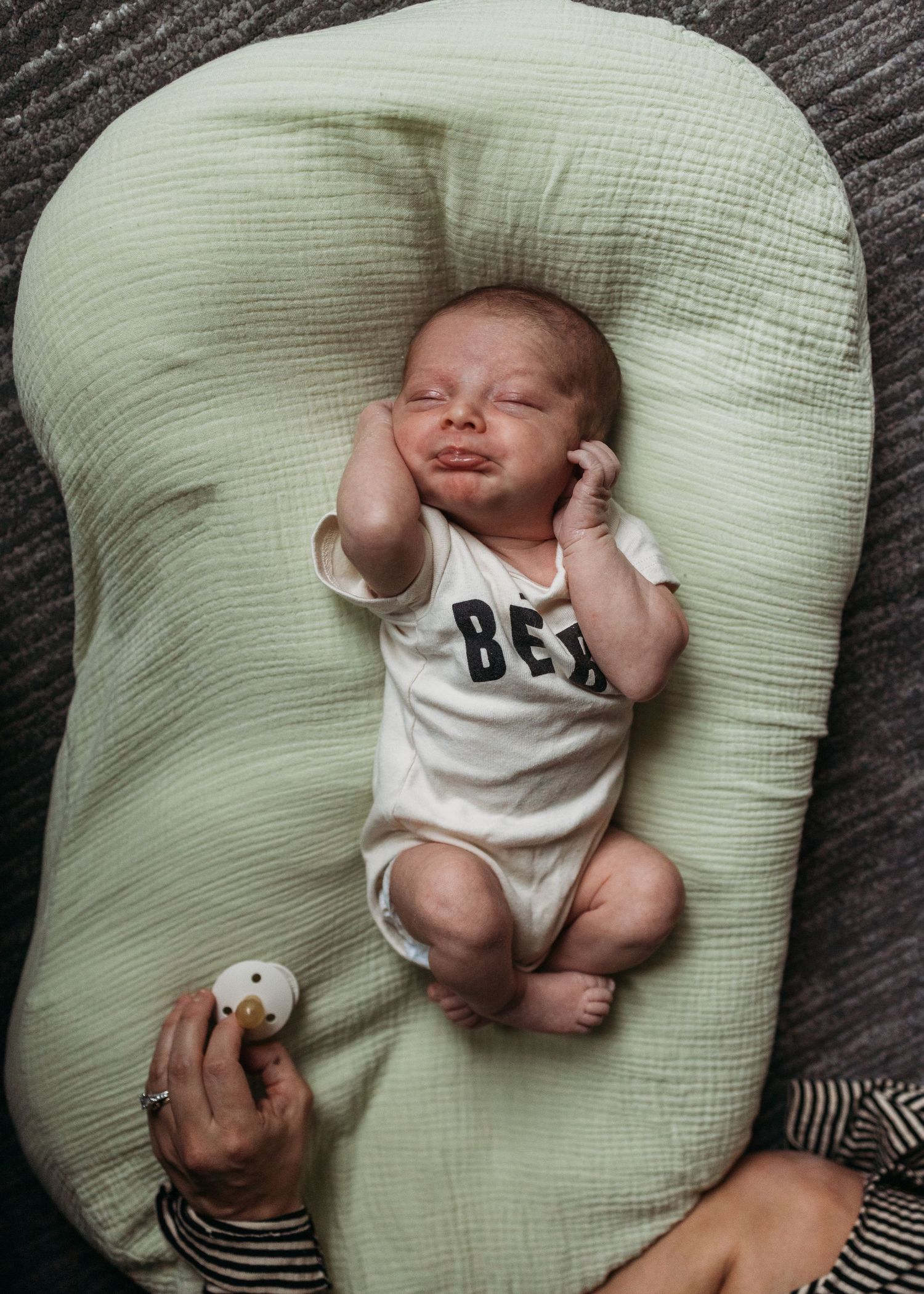 A baby in a white onesie stretches and yawns while lying on a light green nursing pillow.