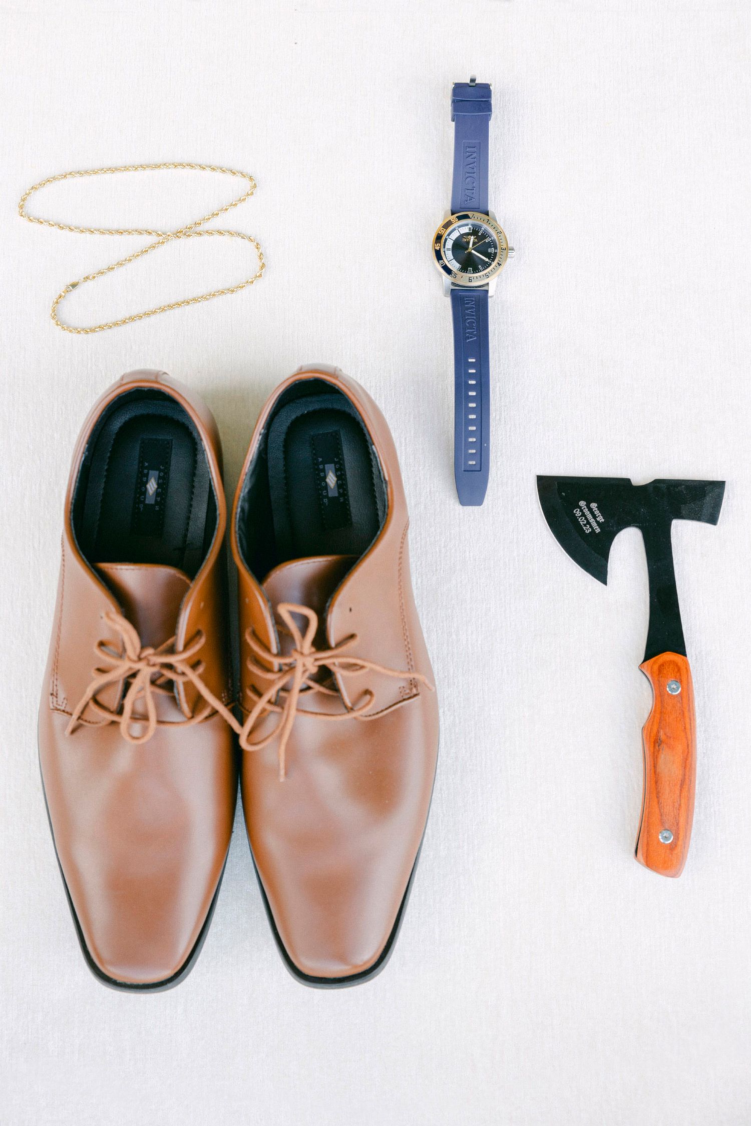 Flat lay of brown leather dress shoes with blue watch and orange-handled axe on white background.