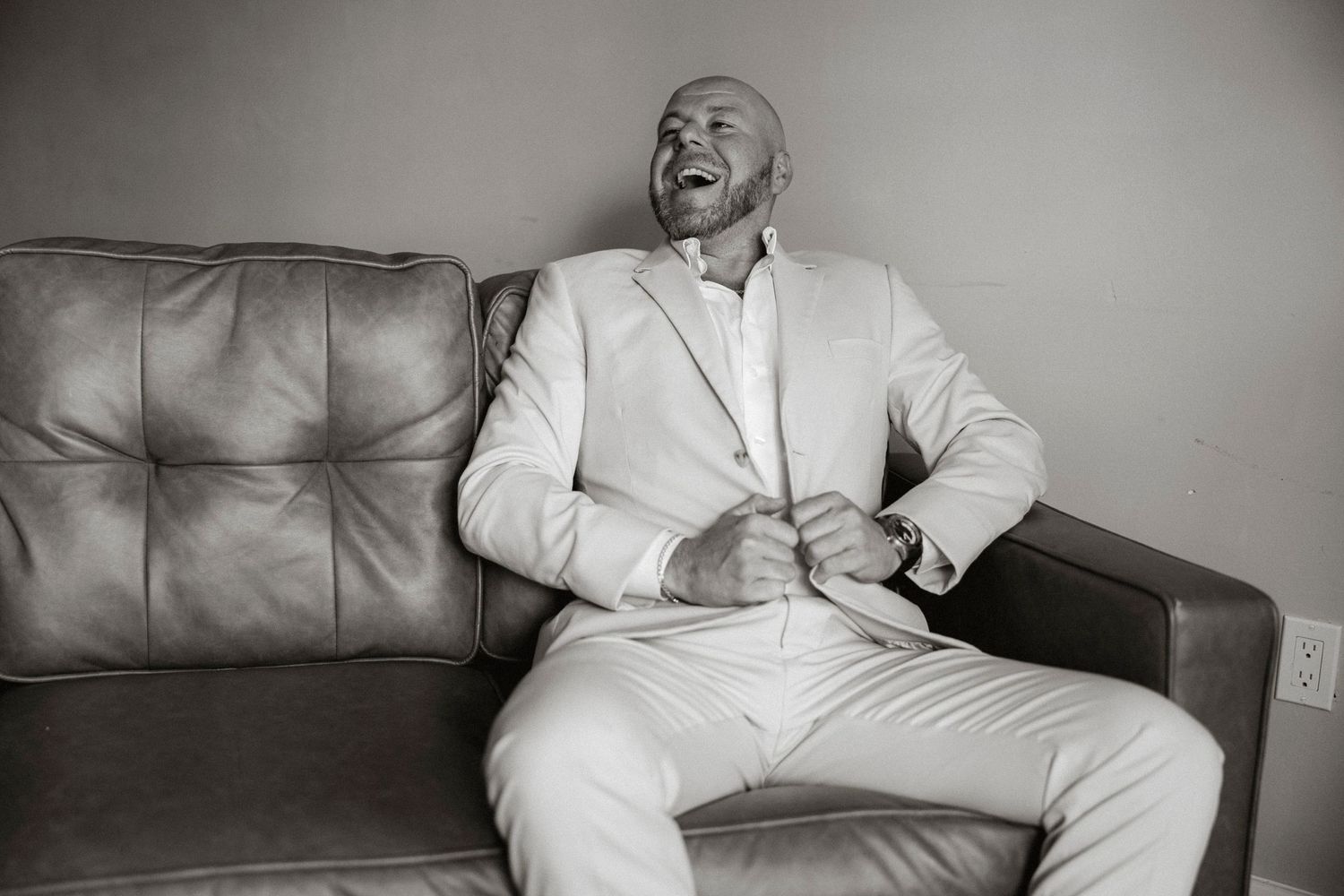 A joyful person wearing an all-white suit sits on a leather couch in a black and white photograph.