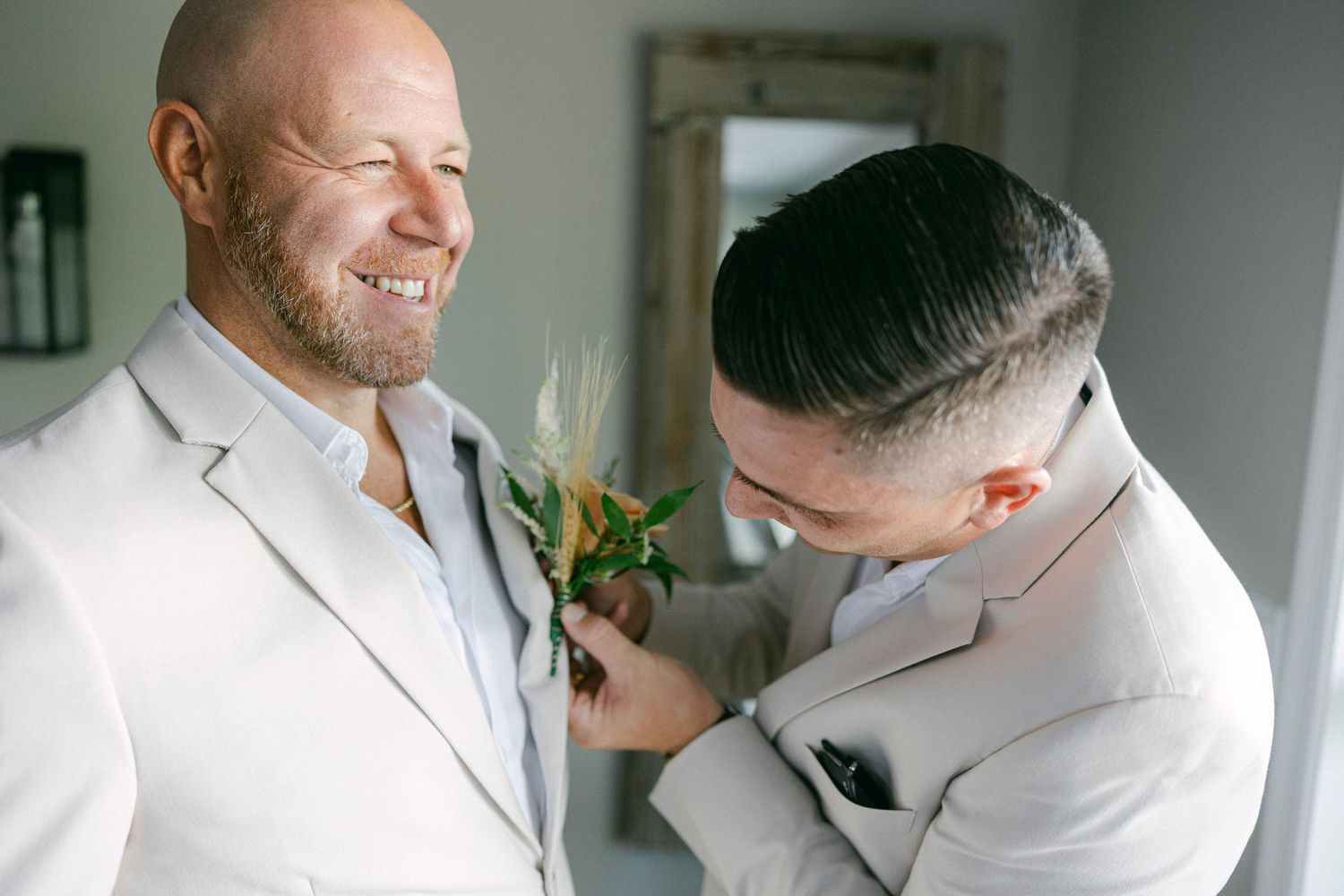 Two people in light colored suits share a joyful moment while adjusting a boutonniere before a formal event.