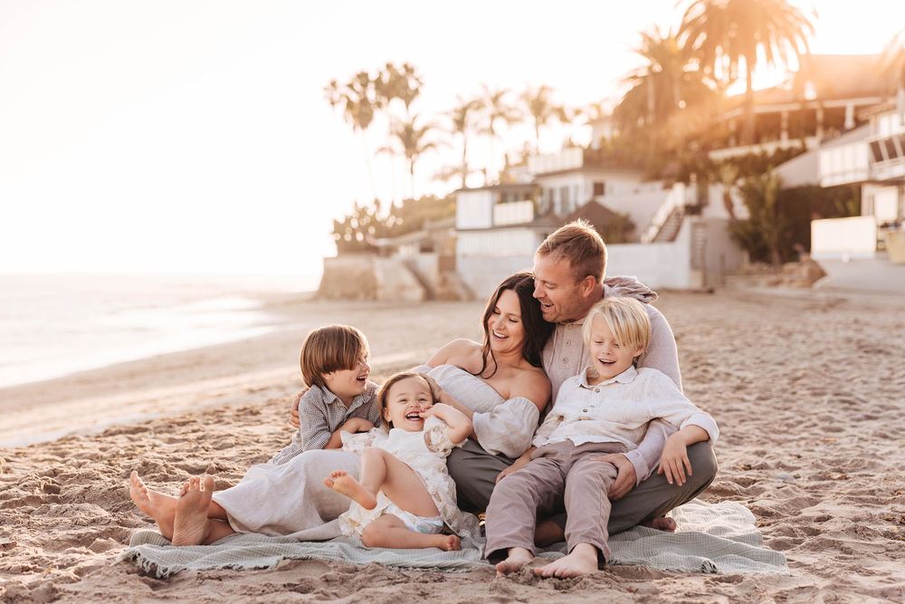 A family relaxes on a blanket at the beach during golden hour with coastal homes visible on the cliffs.