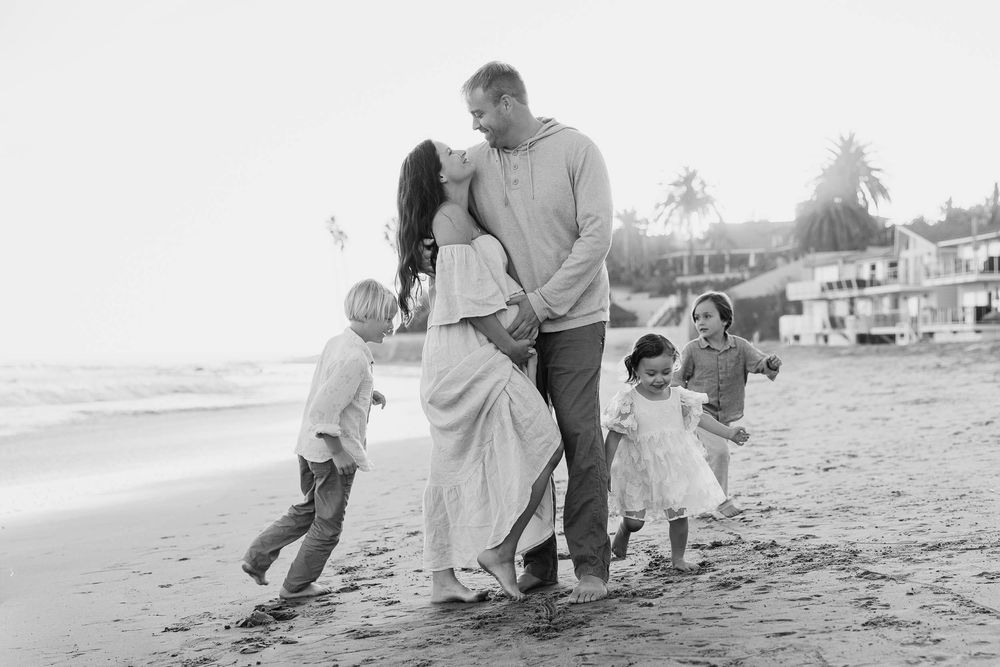 Family plays and embraces on a serene beach with coastal homes in the background.