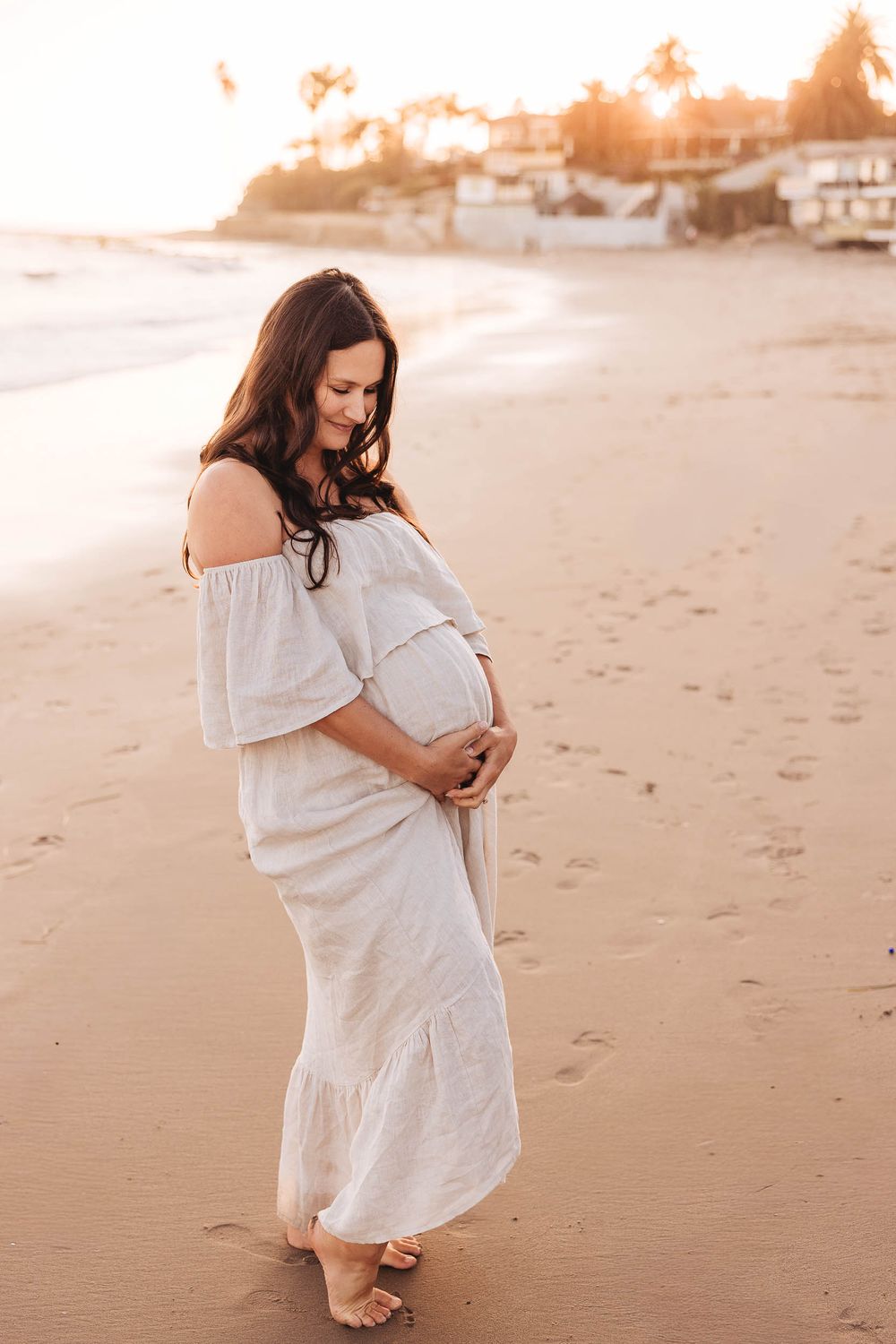 Expectant mother in flowing white dress walks along sandy beach at sunset creating dreamy maternity photos.