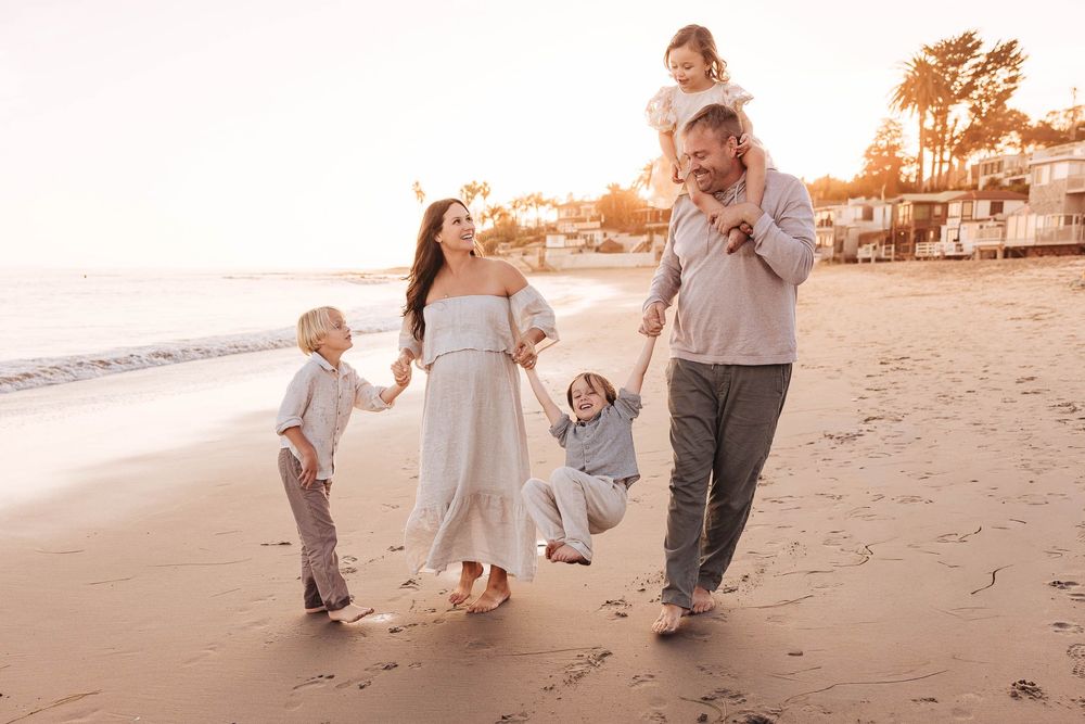 A family walks along the shoreline at sunset with beach houses visible in the background.