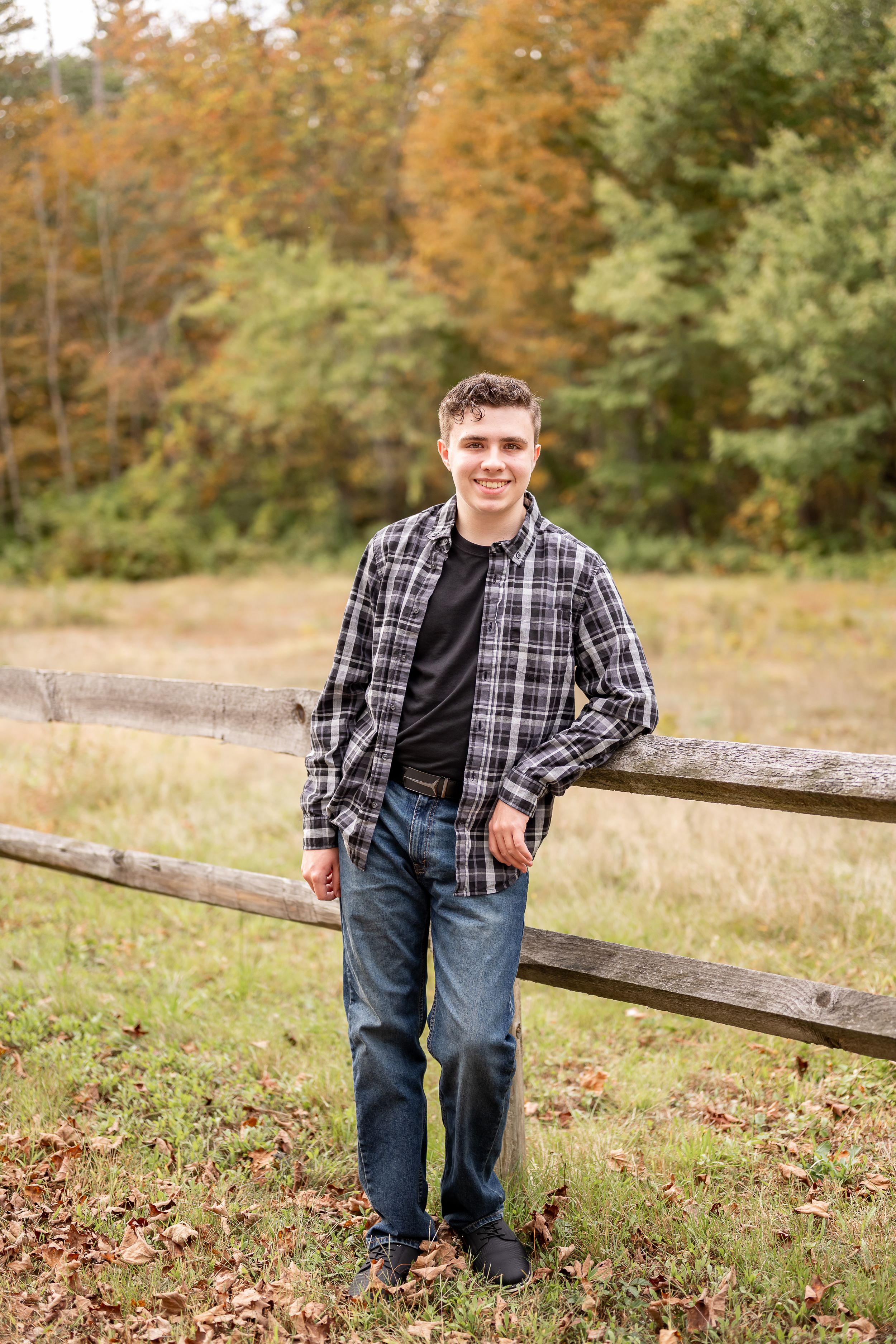 A casual outdoor portrait in autumn featuring someone wearing a plaid shirt and jeans leaning against a wooden fence.