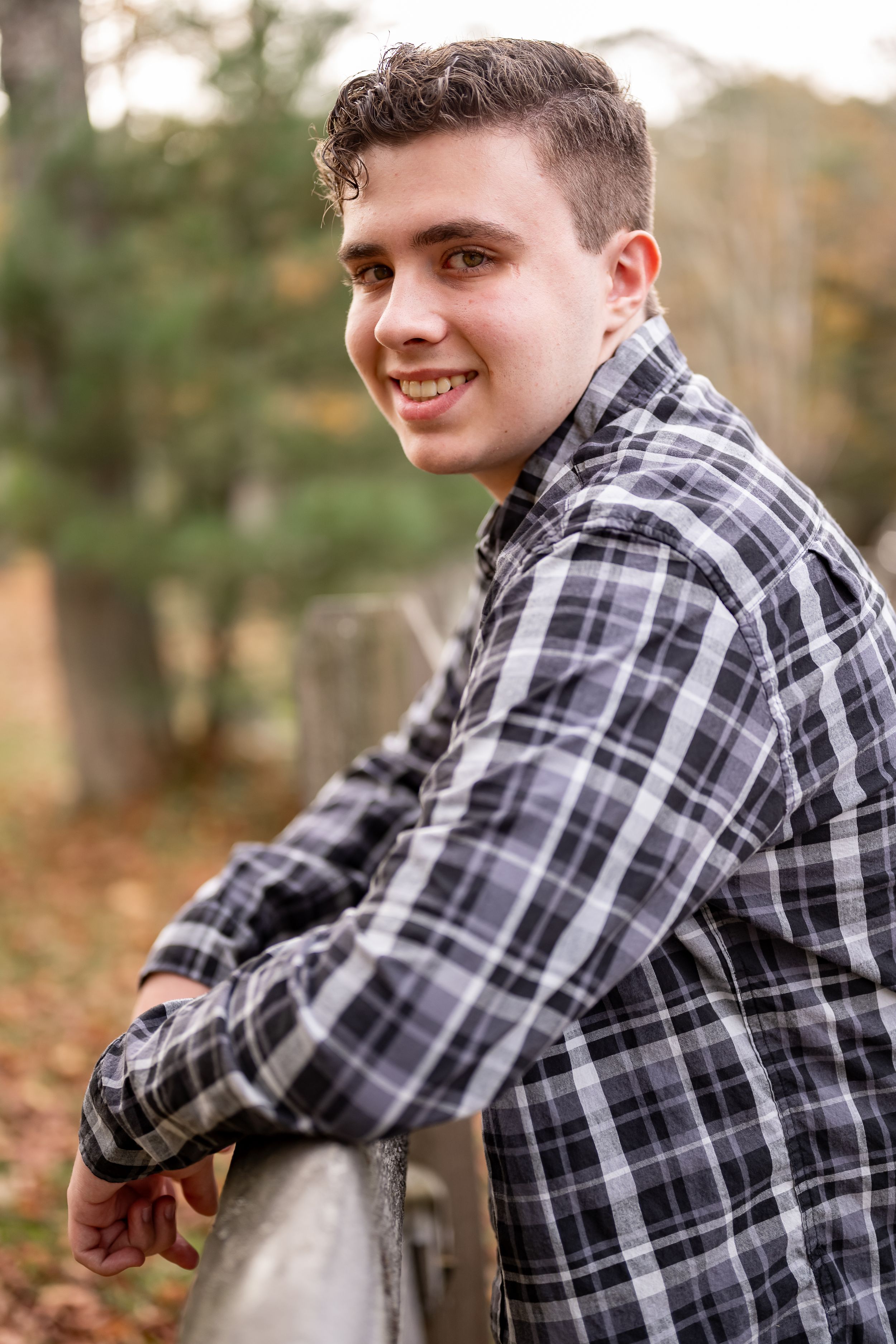 Person in plaid flannel shirt sits casually outdoors in autumn forest setting during photo session.