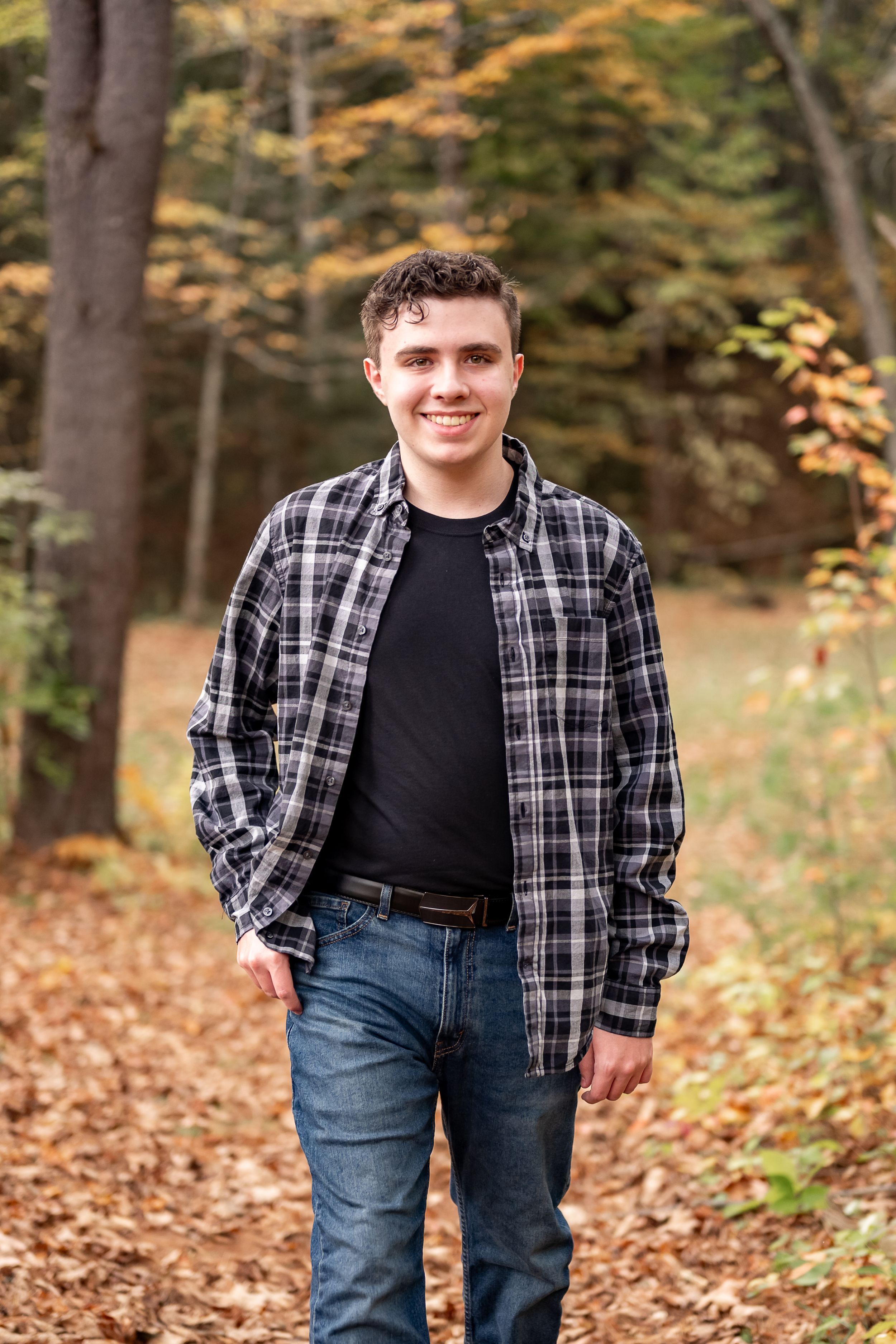 A person in a plaid flannel shirt and jeans poses outdoors among autumn leaves in a wooded setting.