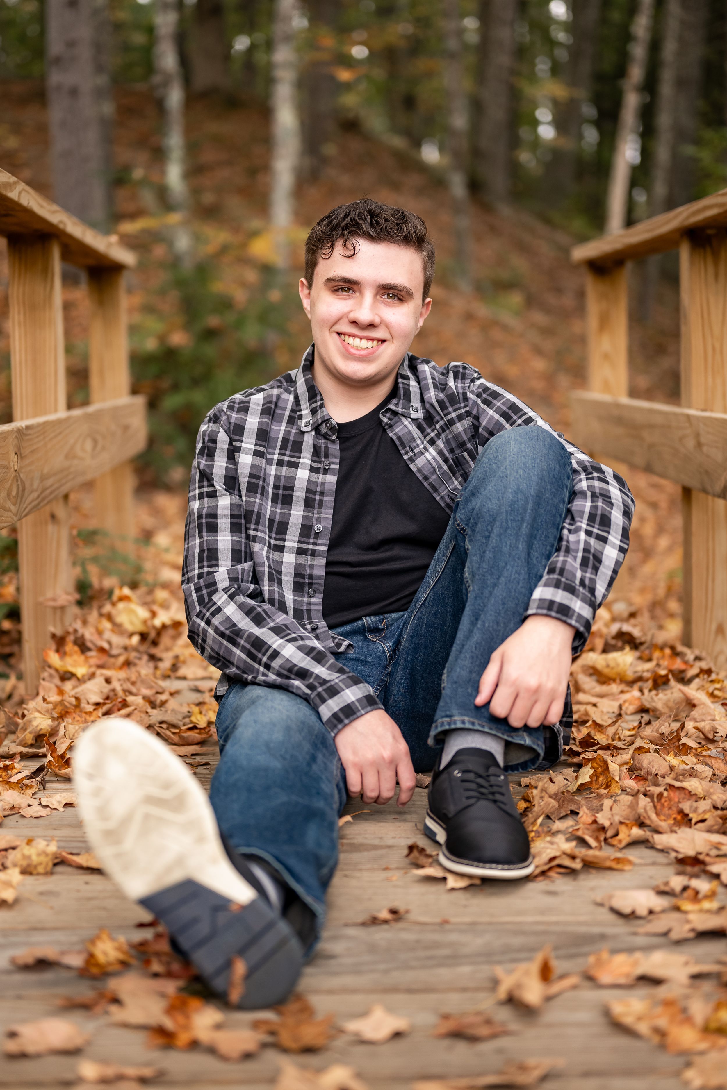 Casual portrait on wooden walkway with autumn leaves wearing plaid shirt and jeans in forest setting.