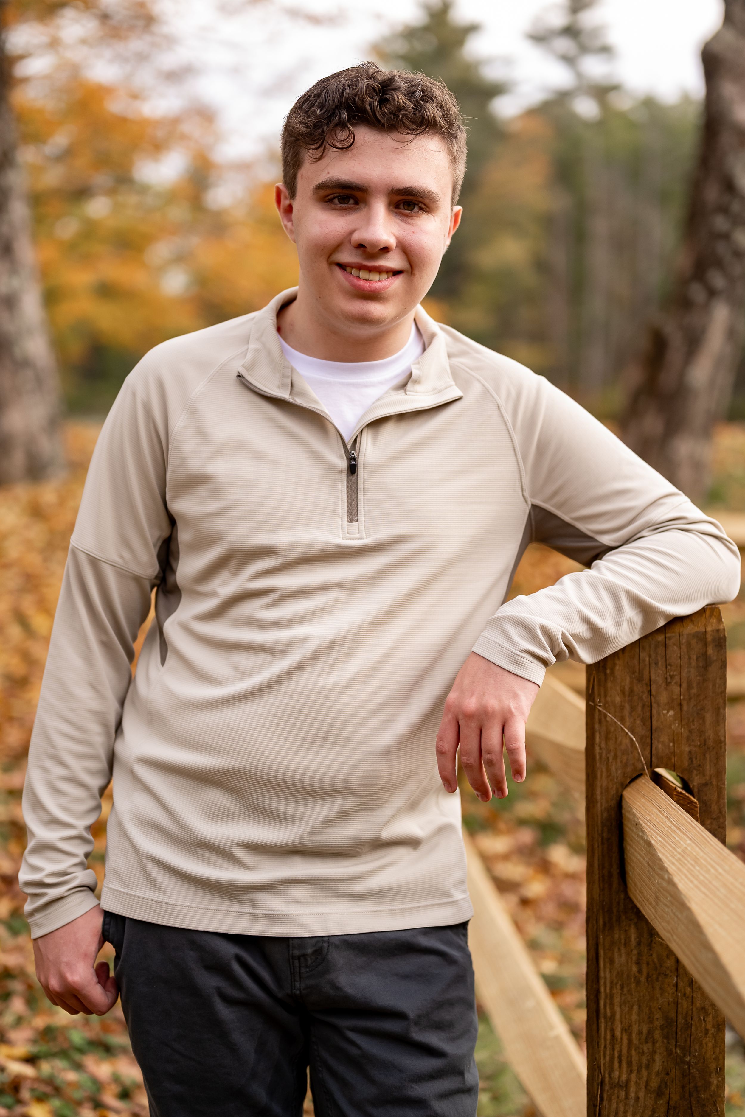 Person in beige quarter-zip pullover poses against wooden railing during autumn with fall foliage in background.