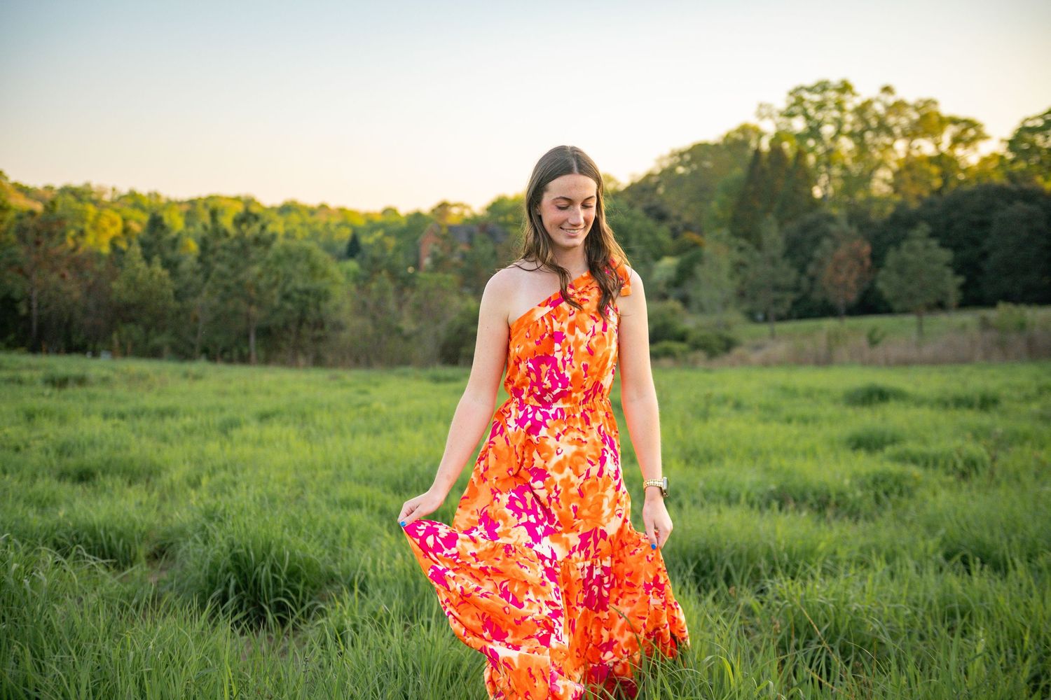 A bright orange and pink floral dress flows in the breeze as someone stands in a sunlit green meadow at golden hour.