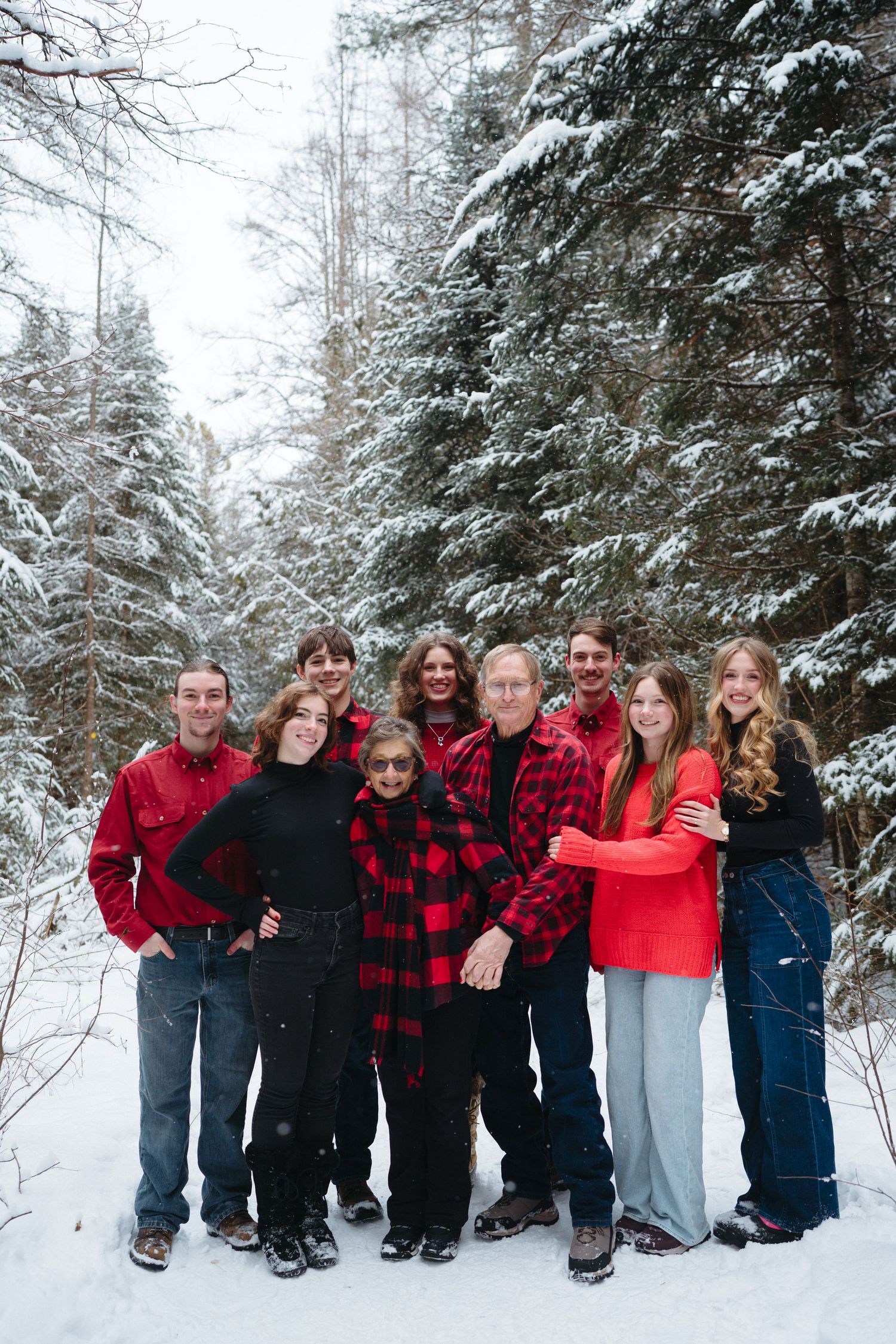 Family members in coordinated red and black outfits pose together in a line against snow-covered pine trees.