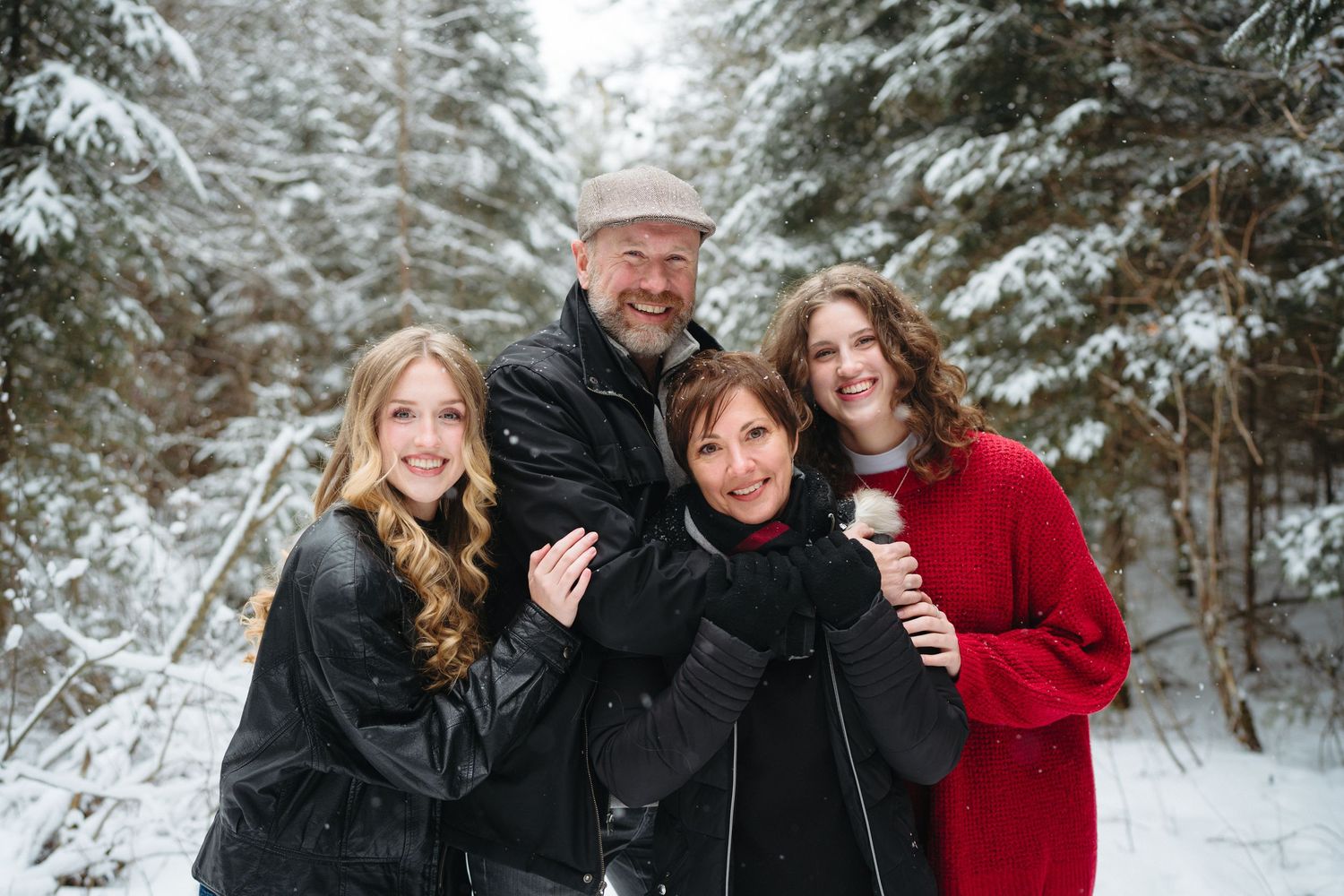 A group stands together smiling outdoors in a snowy winter forest setting with snow-covered evergreen trees in the background.