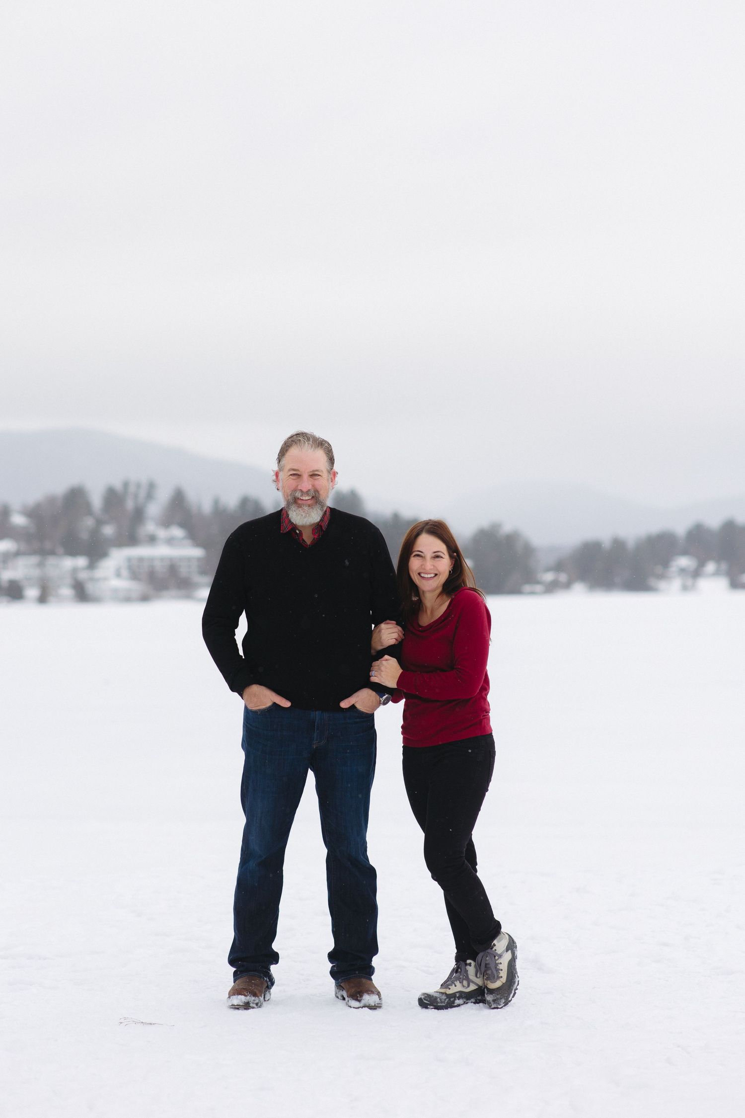 A couple stands together on a snowy landscape with mountains and trees in the background.
