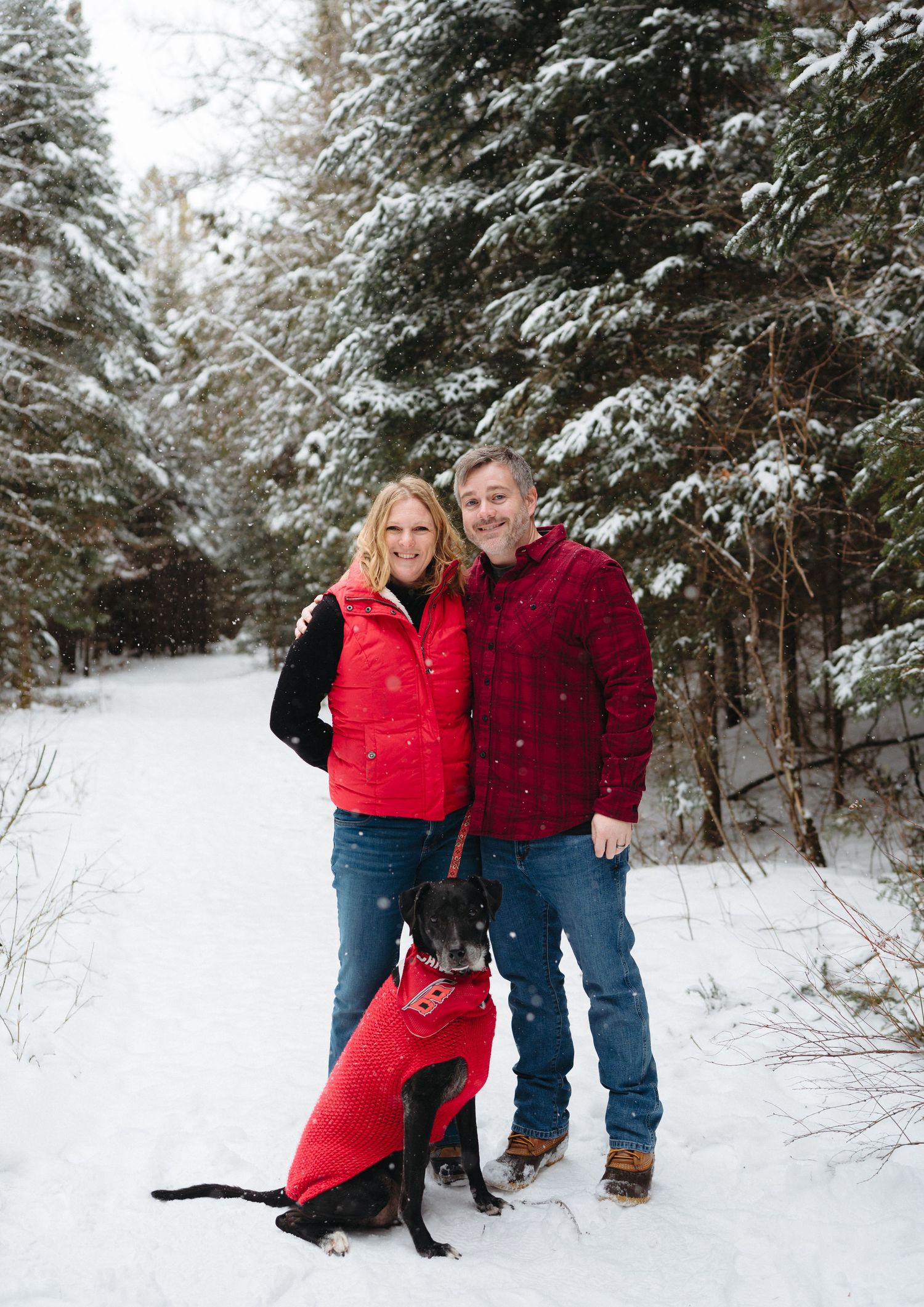 A couple and their black dog wearing a red coat pose together on a snowy winter trail surrounded by evergreen trees.