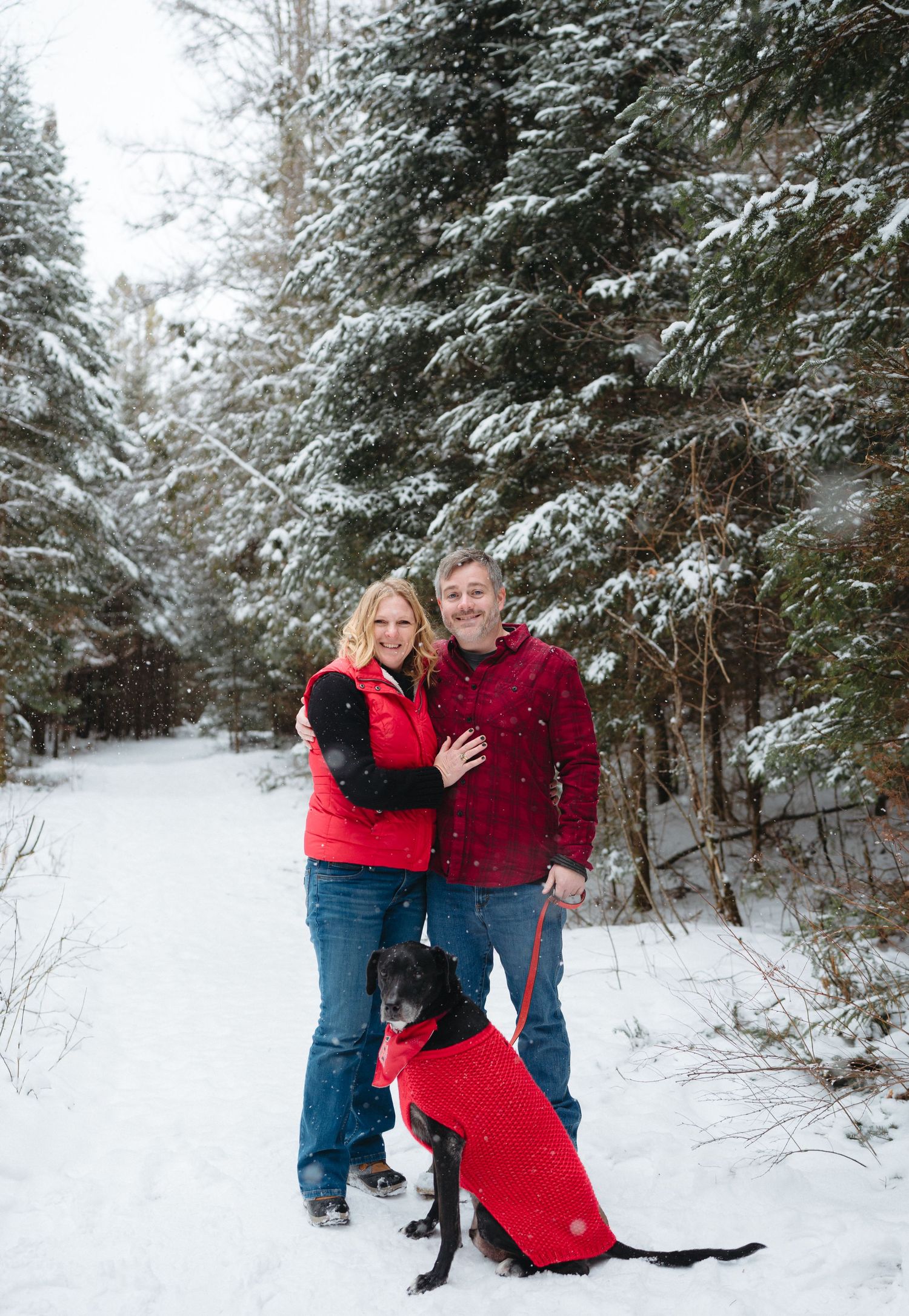 A couple stands with their black dog wearing a red jacket on a snowy forest path among snow-covered evergreens.
