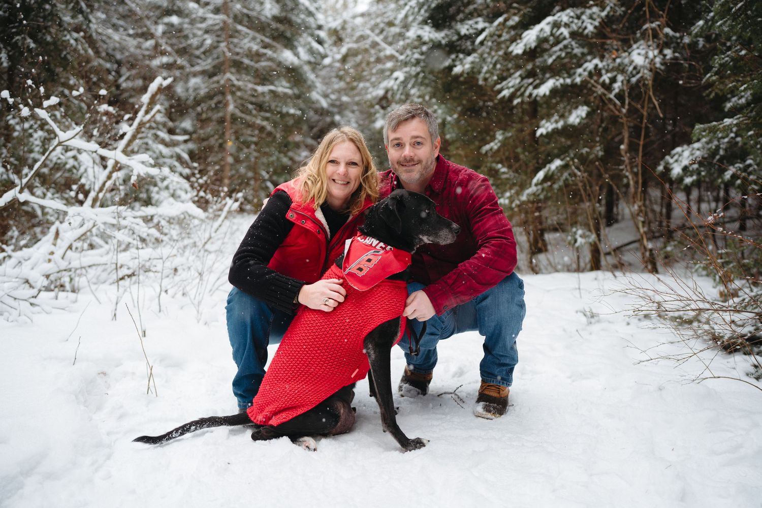 Two people in winter coats pose with their black dog wearing a red coat on a snowy forest trail.