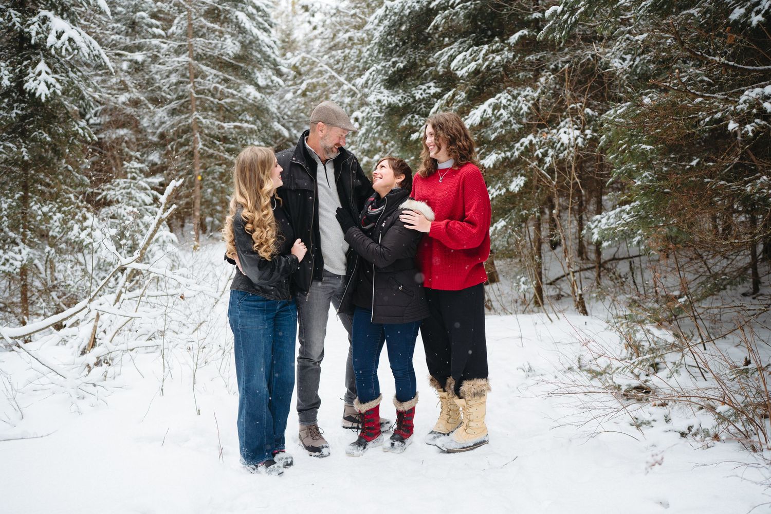 A group of people laugh together while standing in a snowy forest setting wearing winter coats.